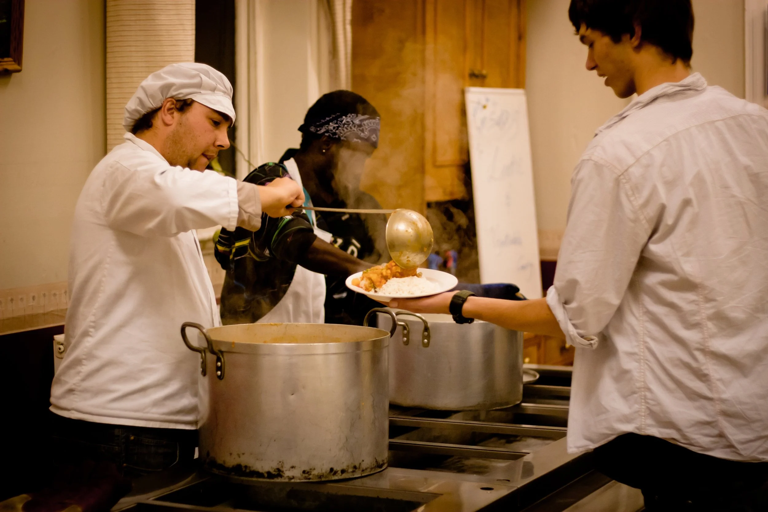Four people serving food from large pots at a buffet or self-serve station, with one person in a chef's hat, and another handing a plate of food.