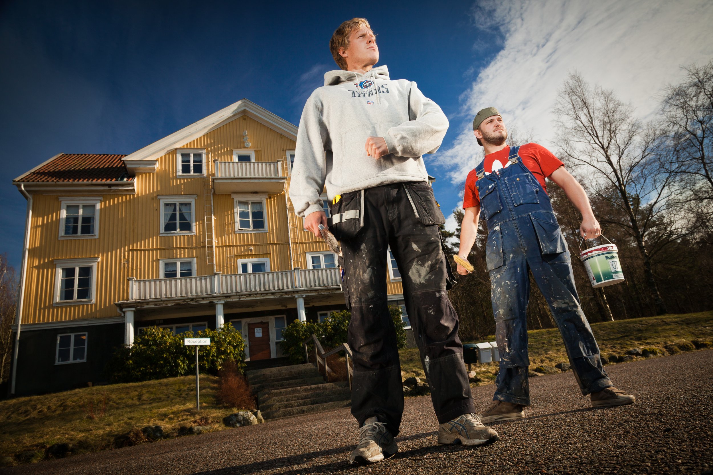Two construction workers standing in front of a yellow house. One holding a paintbrush and the other holding a paint bucket, both dressed in work clothes with paint marks.