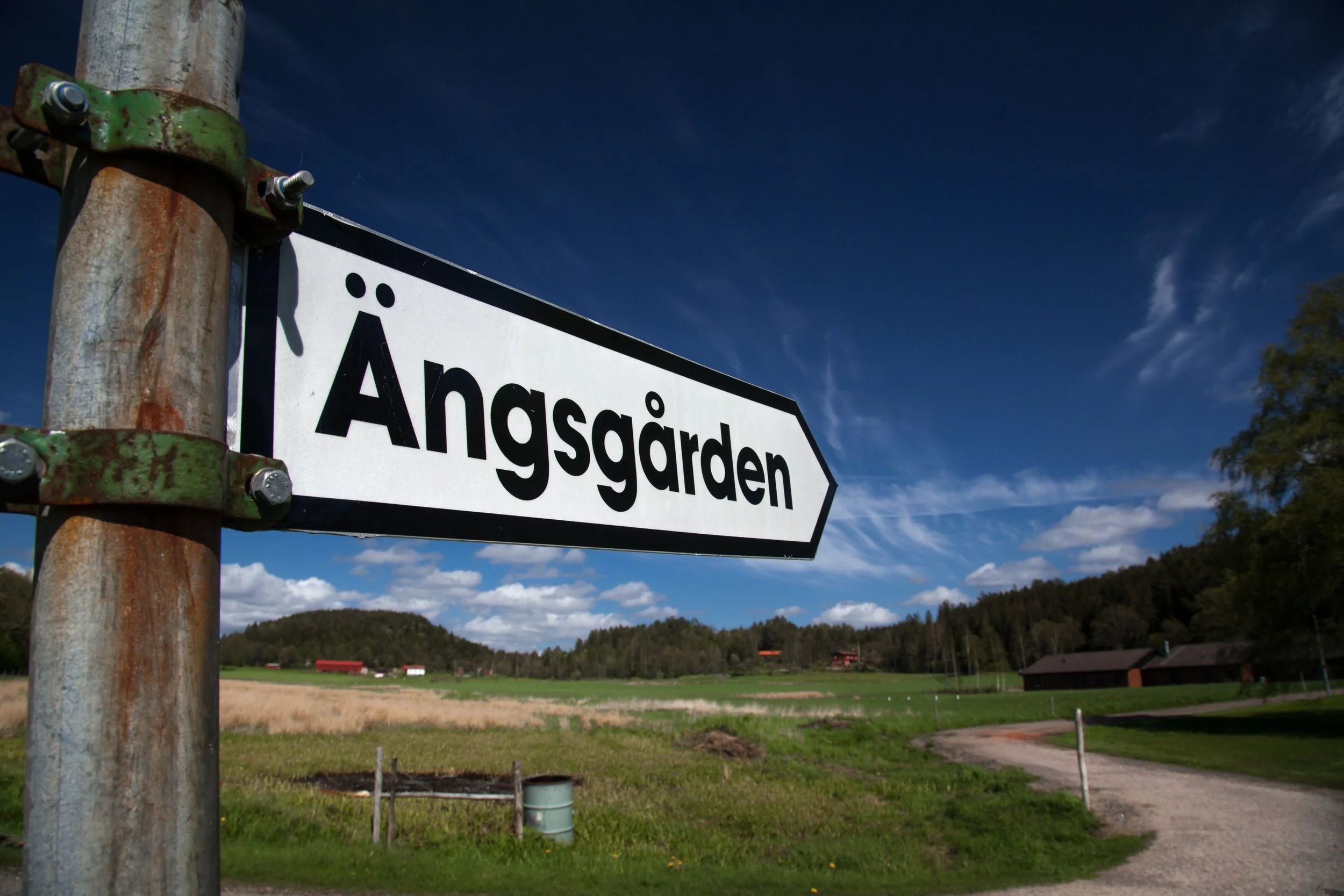 A directional road sign reading 'Ånggården' pointing towards a rural area with fields, scattered buildings, and a wooded hillside under a blue sky with some clouds.