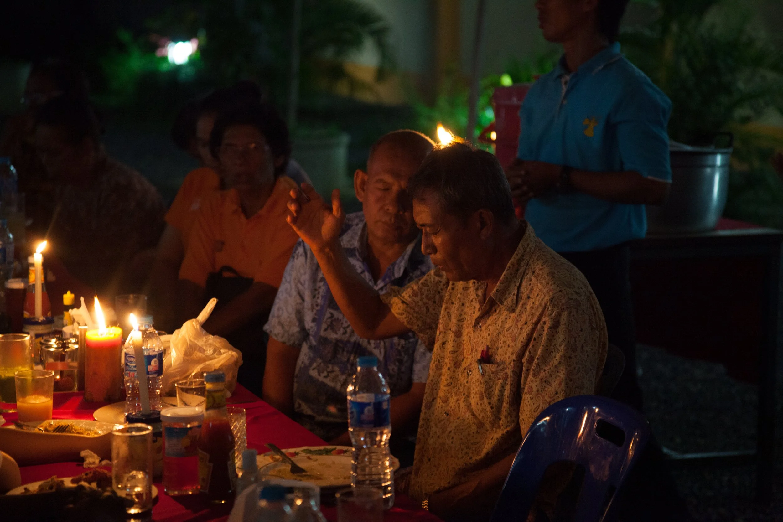People gathered around a table with candles and food during a nighttime outdoor event.