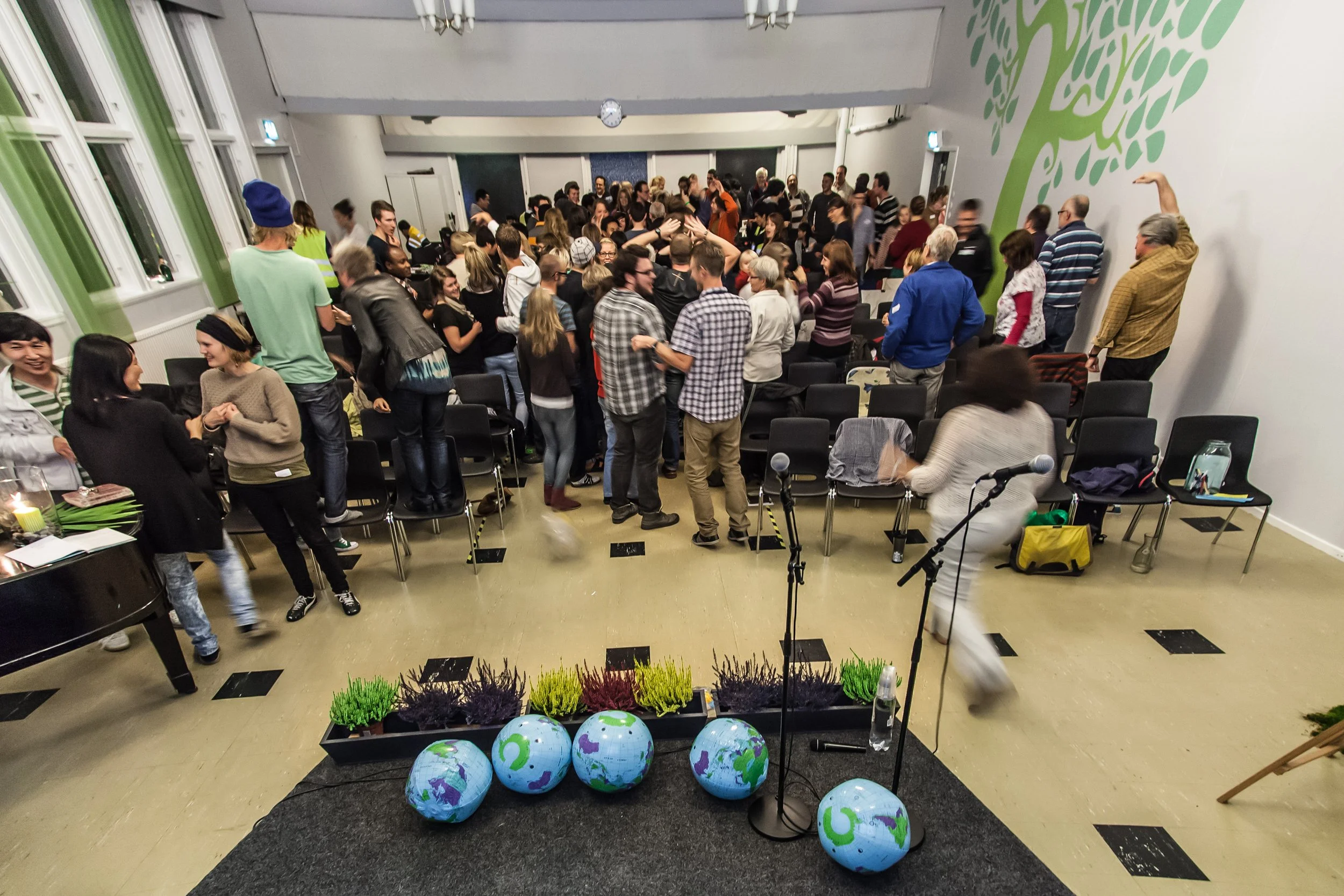 People gathered in a large room with chairs and microphones, likely for an event or presentation, with some people talking and socializing.
