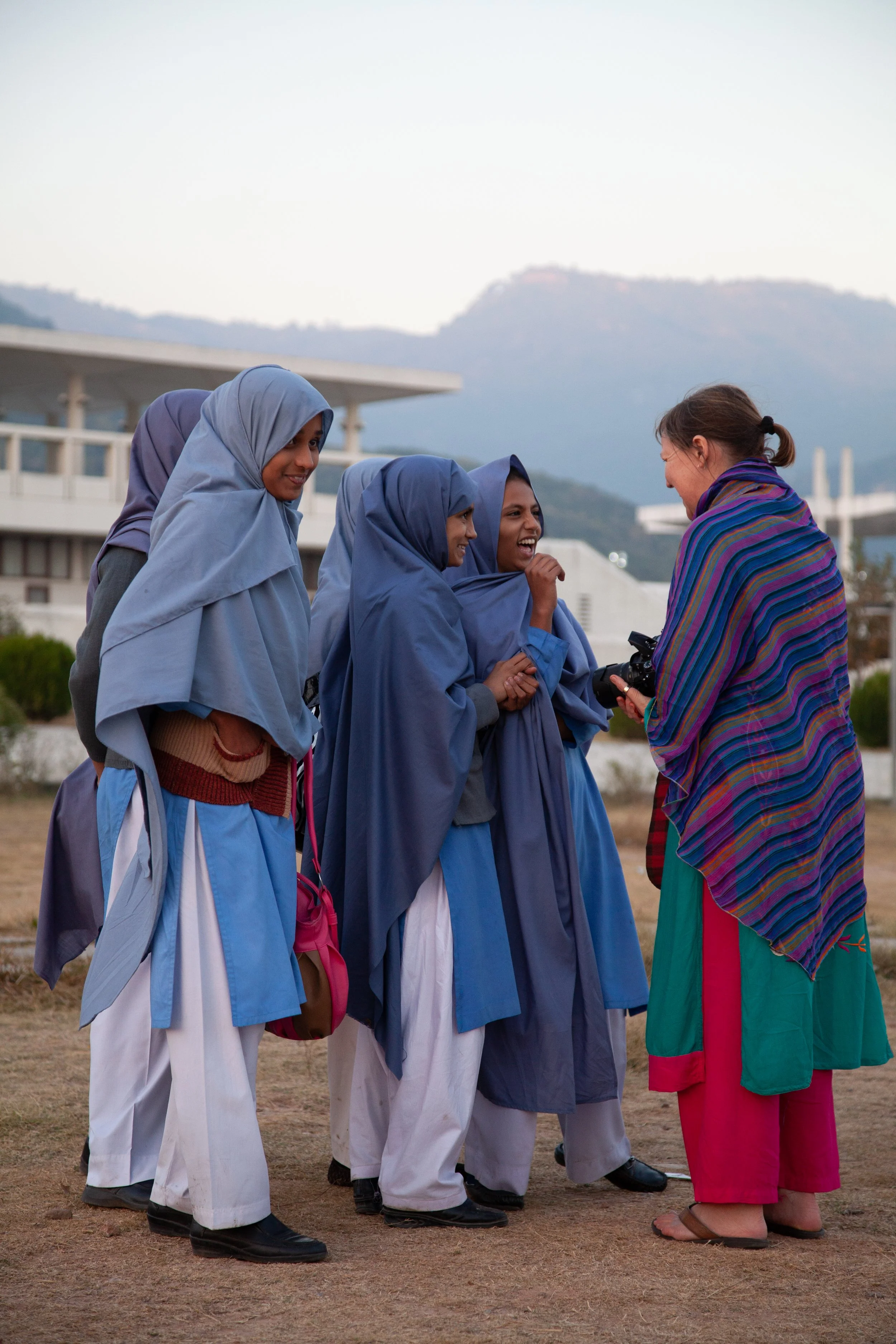 A woman with a camera talking to a group of women and girls wearing blue traditional clothing and headscarves outdoors, smiling and engaging in conversation.