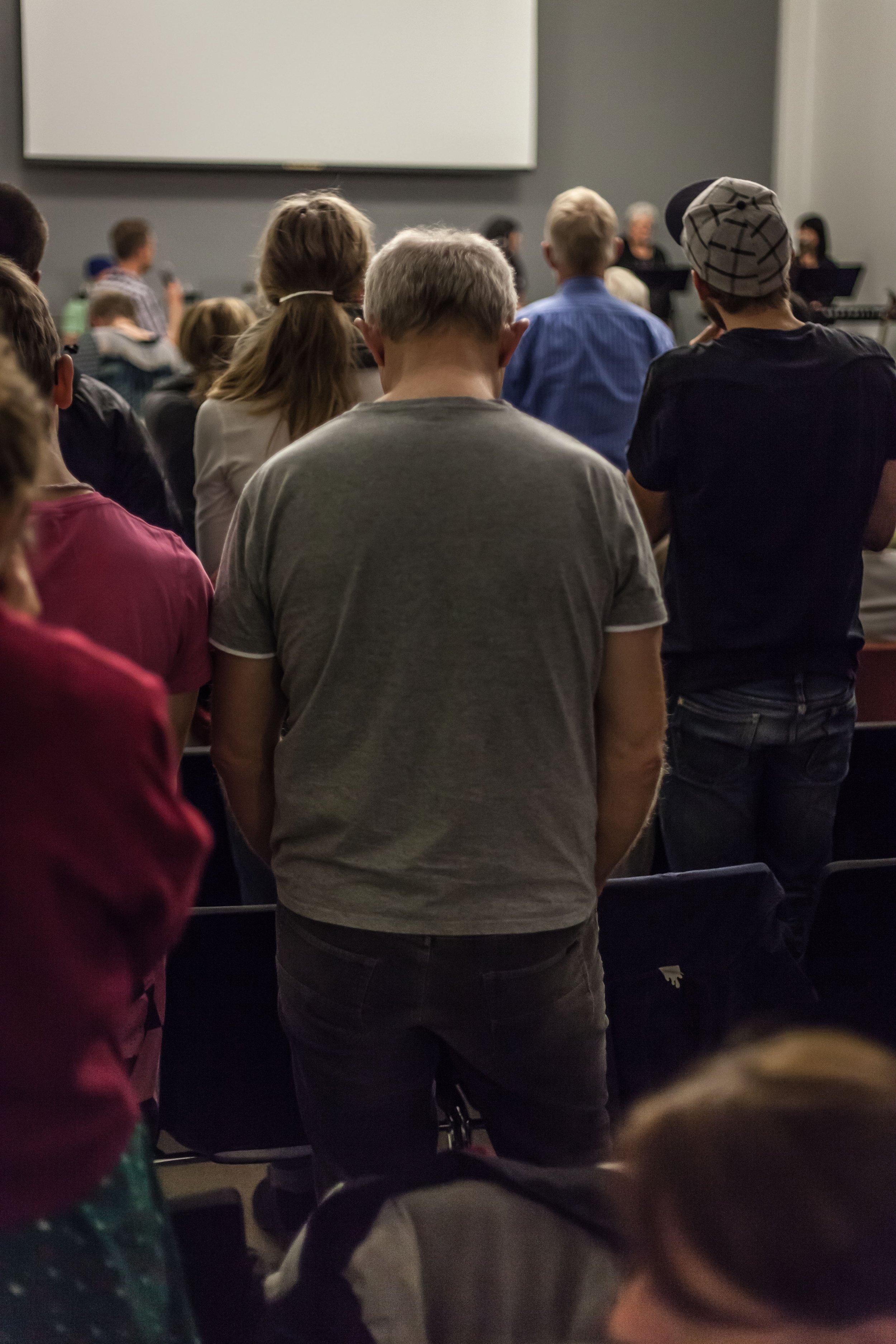 A group of people standing in a room, facing a stage or screen at the front, possibly during a presentation or event.