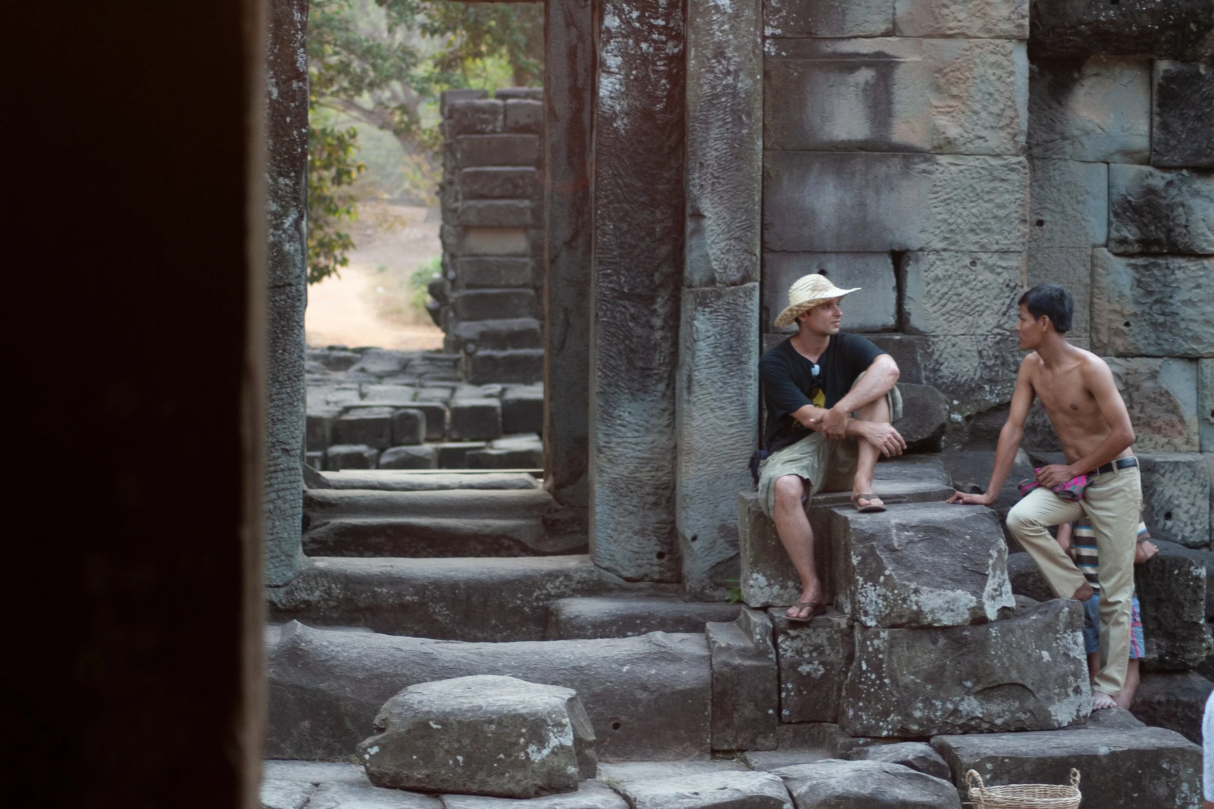 Two young men converse on ancient stone ruins, one shirtless and the other wearing a straw hat surrounded by large stone blocks and steps.