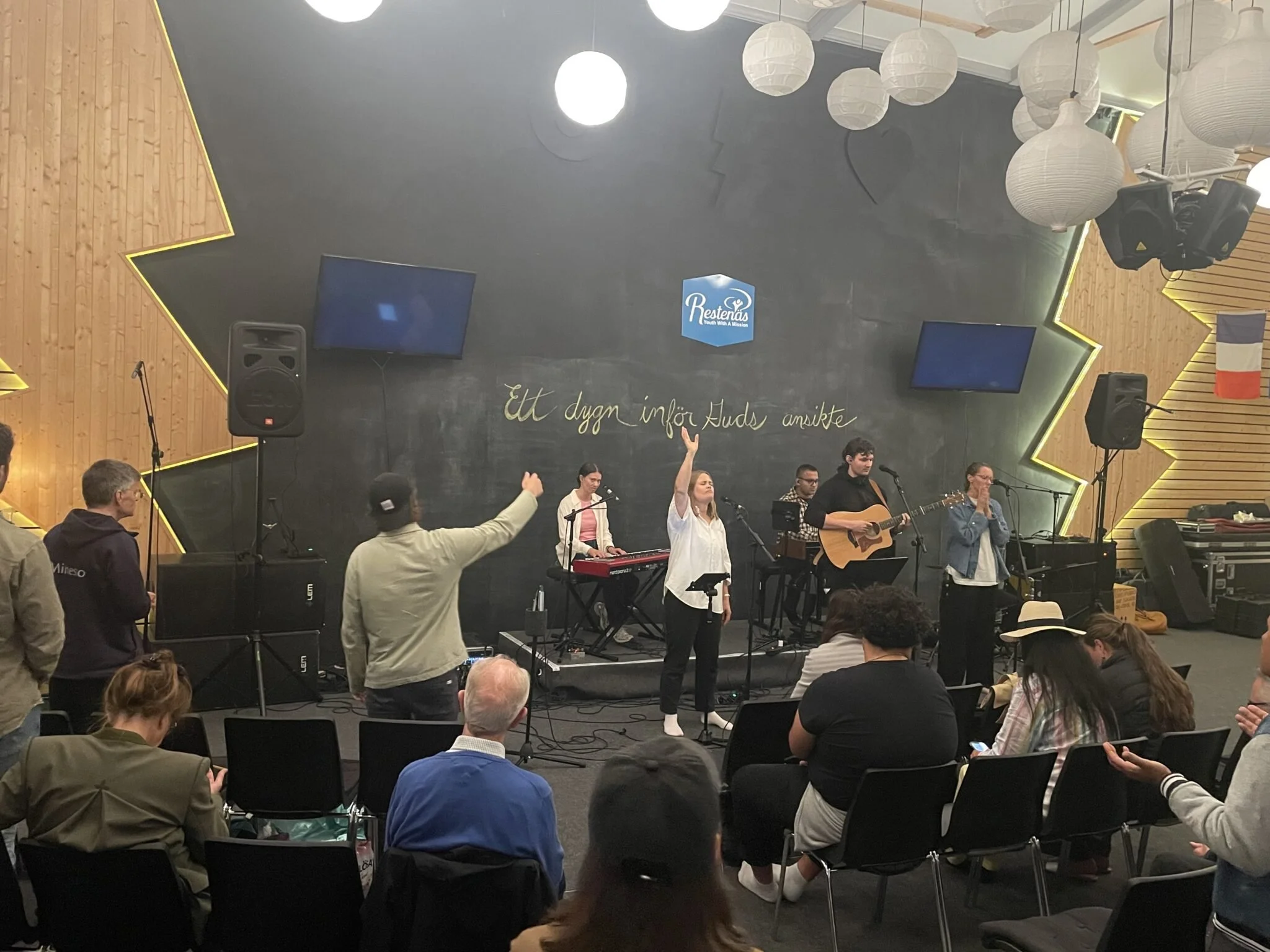 A group of performers on stage singing and playing instruments, with audience members seated and watching. The stage has a black chalkboard with gold writing, and the background features geometric wooden panels with yellow lighting accents. There are hanging white lanterns and two large screens above the stage.