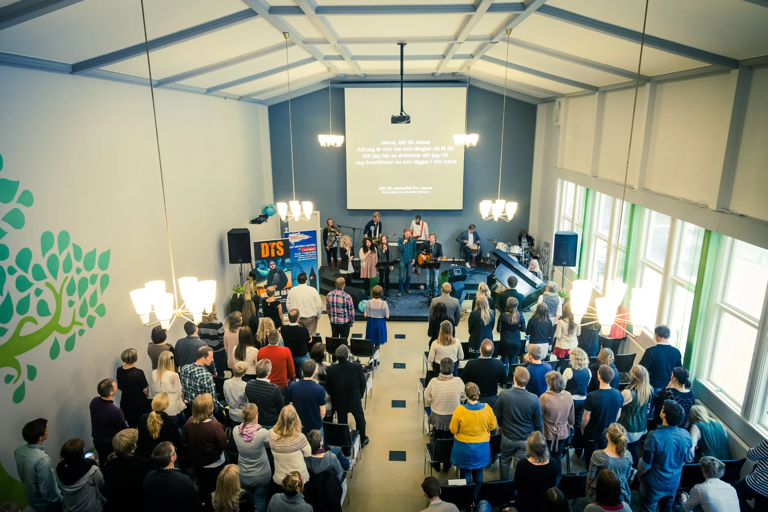 A congregation gathered in a church or hall for a worship service or concert. The congregation is facing a stage where musicians and singers are performing. Large windows on the side let in natural light. There is a large screen displaying lyrics in 