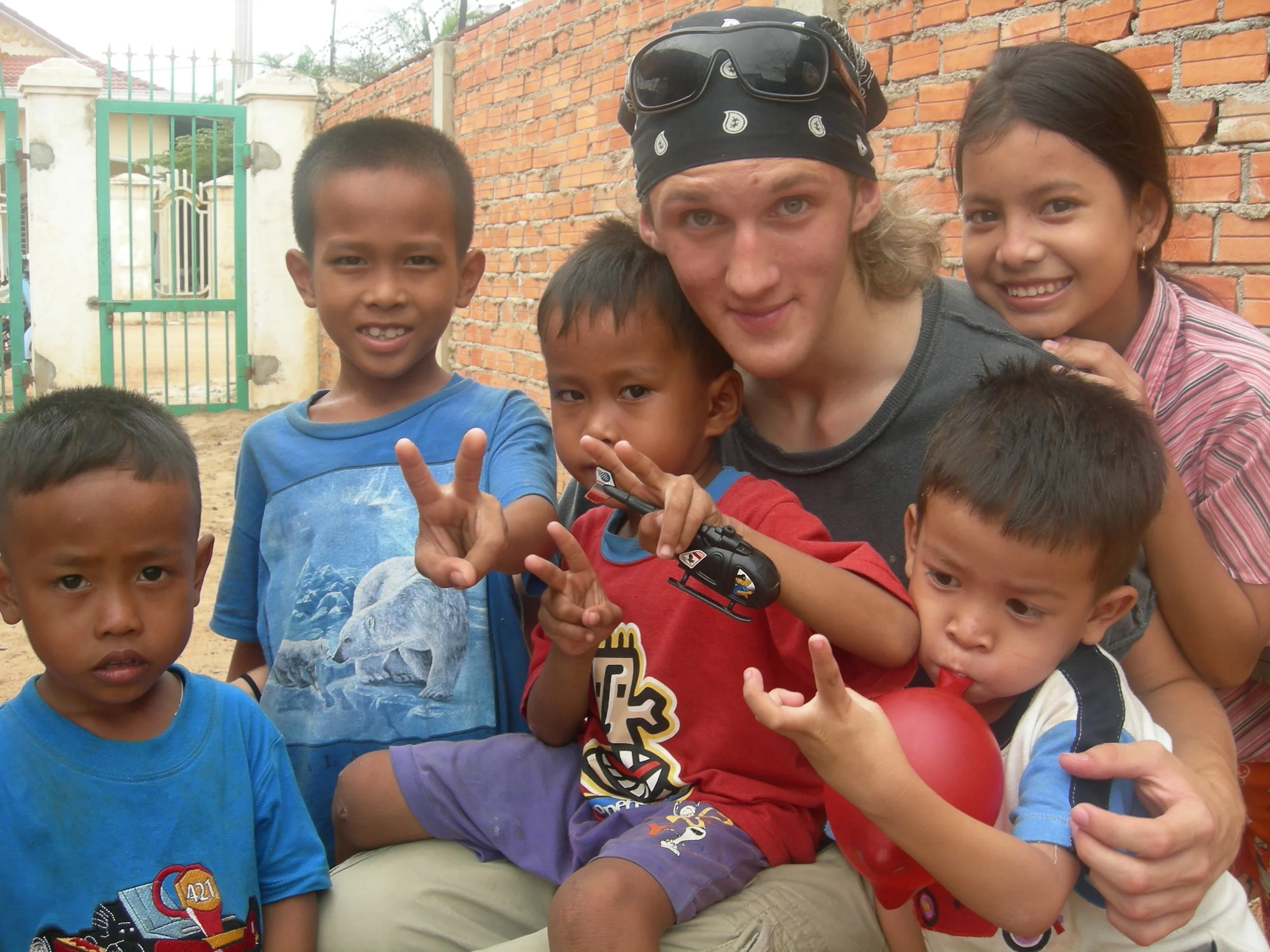 A group of children and a young man are gathered outdoors in front of a brick wall, smiling and posing for the camera. The children are of varying ages and are wearing casual clothing.