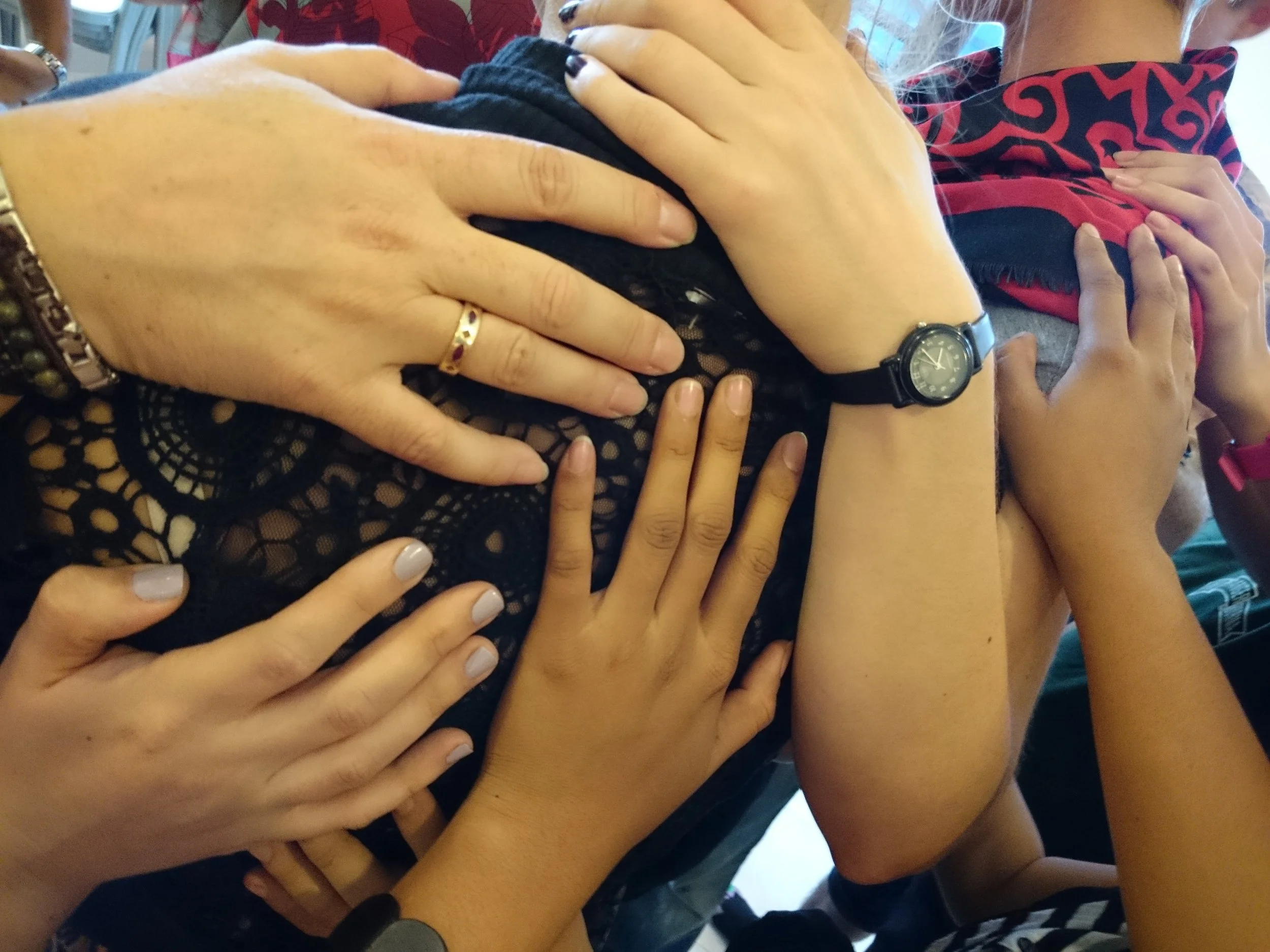 Multiple hands, belonging to different people, are overlapping on a person's shoulder and chest area. The person is wearing a black lace top and a wristwatch. One hand is wearing a ring, and others have painted nails.