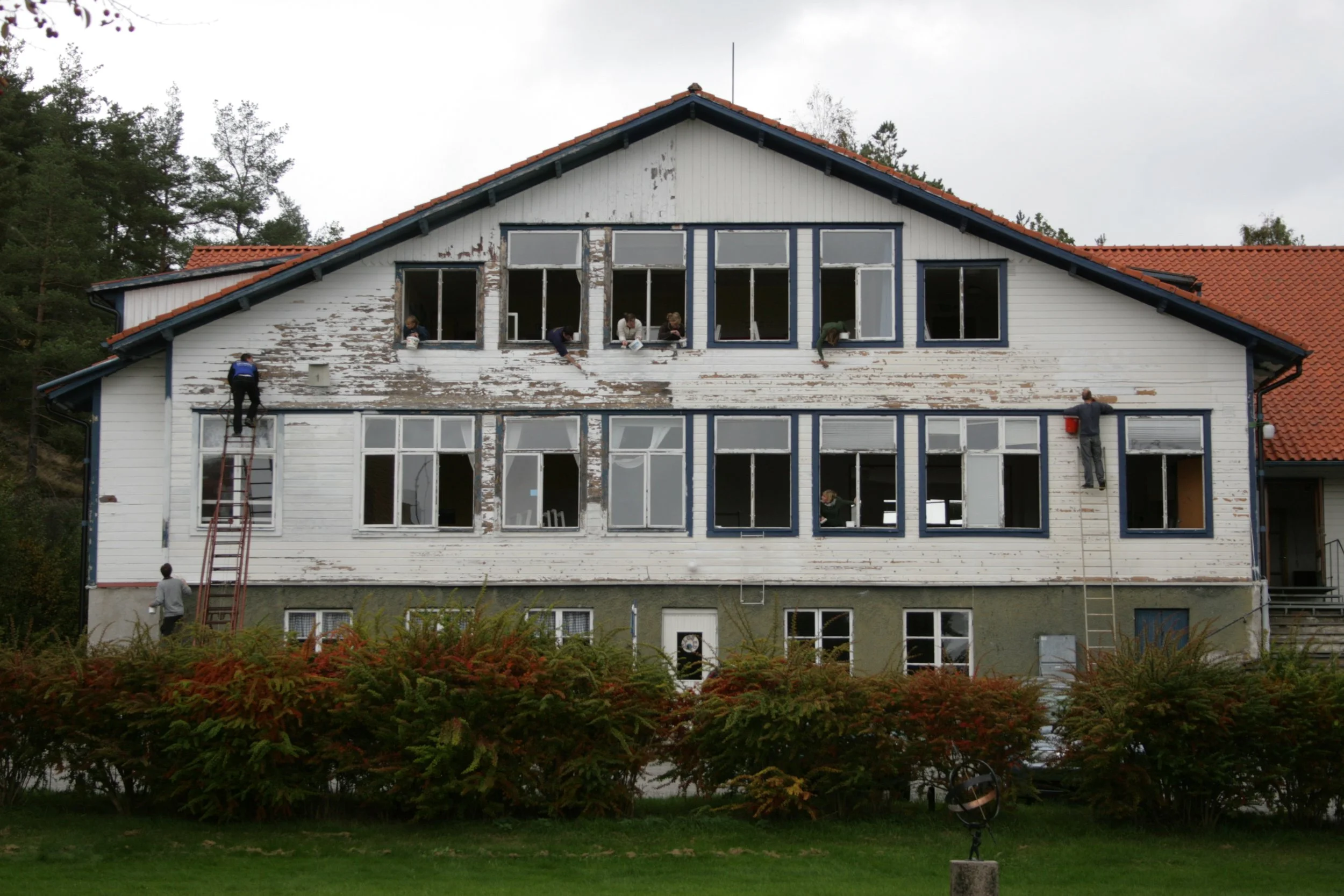 People renovating the exterior of a multi-story house, painting and scraping paint from the wooden siding.