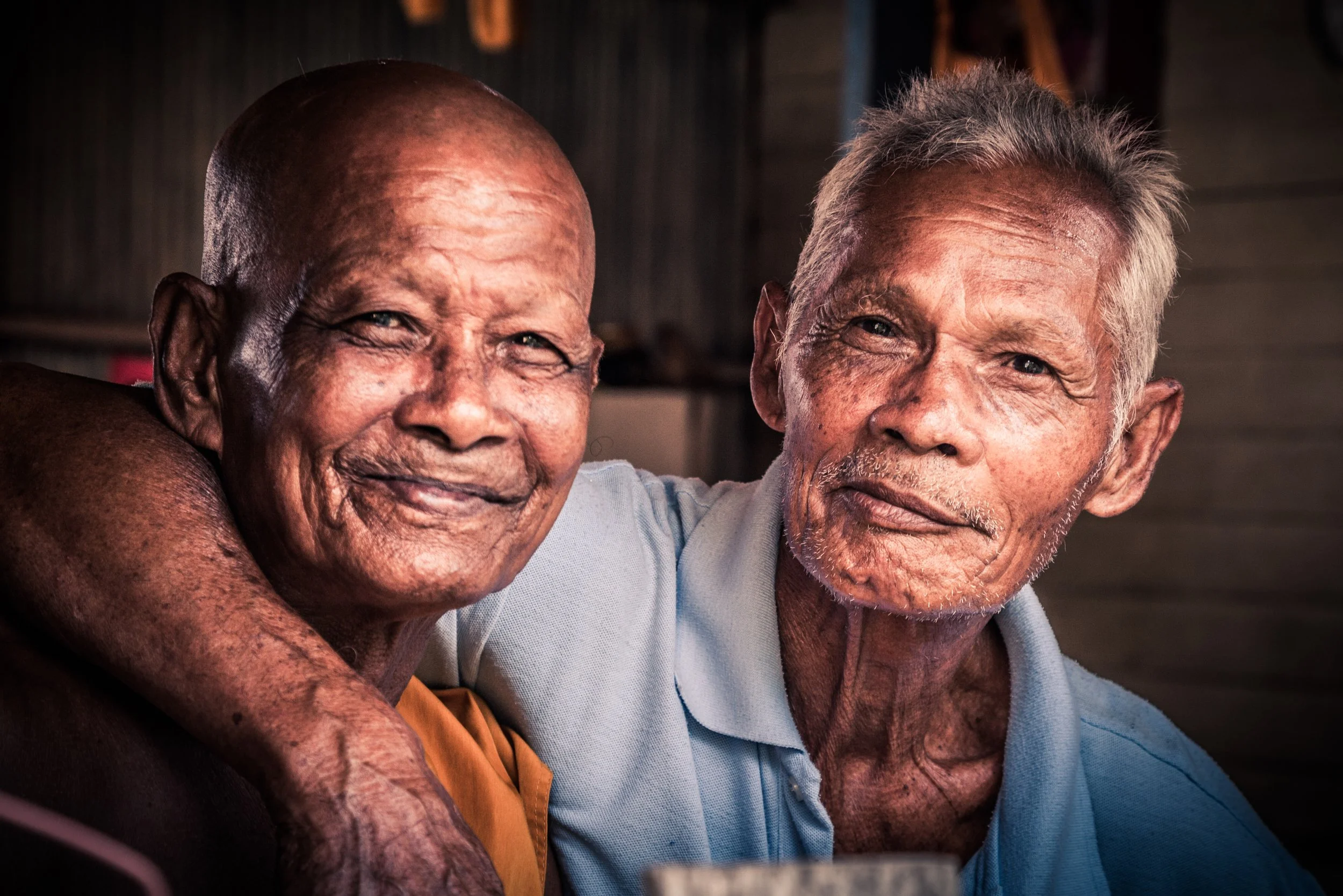 Two elderly men with gray hair and smiling faces sitting close together, one with his arm around the other, in a warmly lit indoor setting.