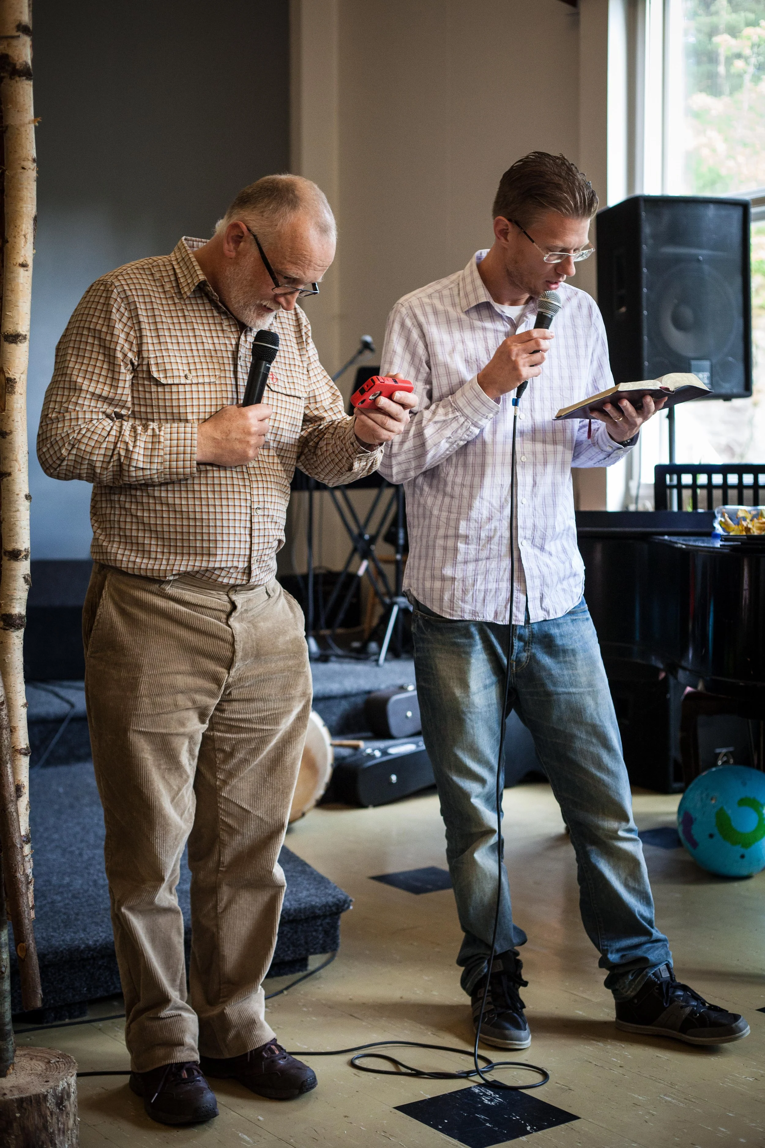 Two men praying with microphones, one reading from a book, in a room with musical equipment and a window.
