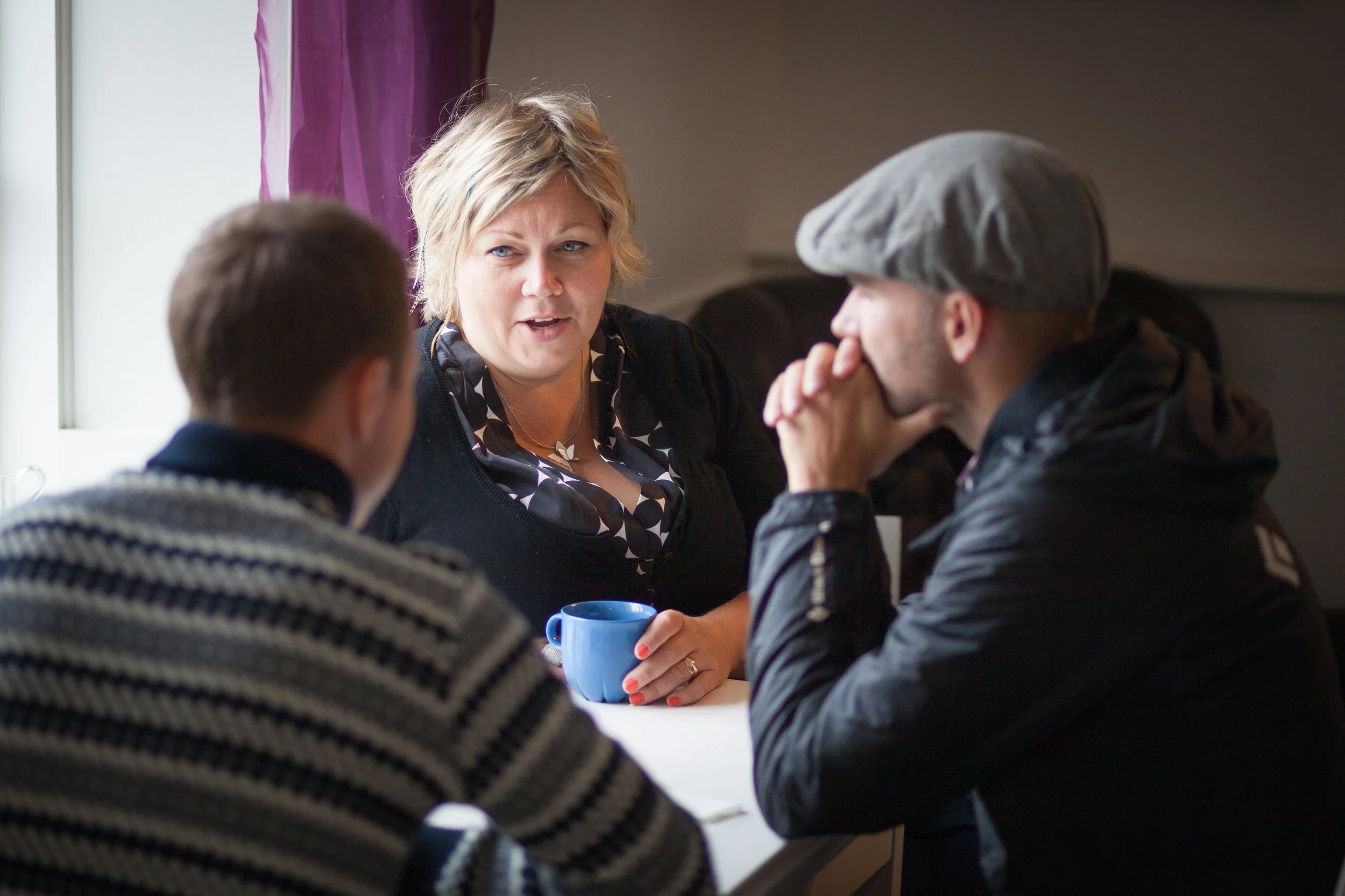 A woman with blonde hair talking to two men at a table, one wearing a gray hat and dark clothing, the other with short dark hair and striped sweater, in a room with a window and purple curtains.
