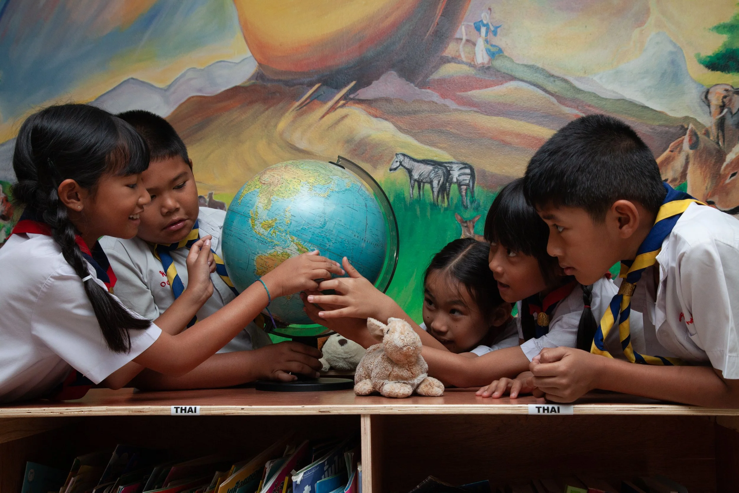 Group of children in scout uniforms gathered around a globe, examining it together, with a colorful mountain landscape mural in the background and a stuffed rabbit on the table.