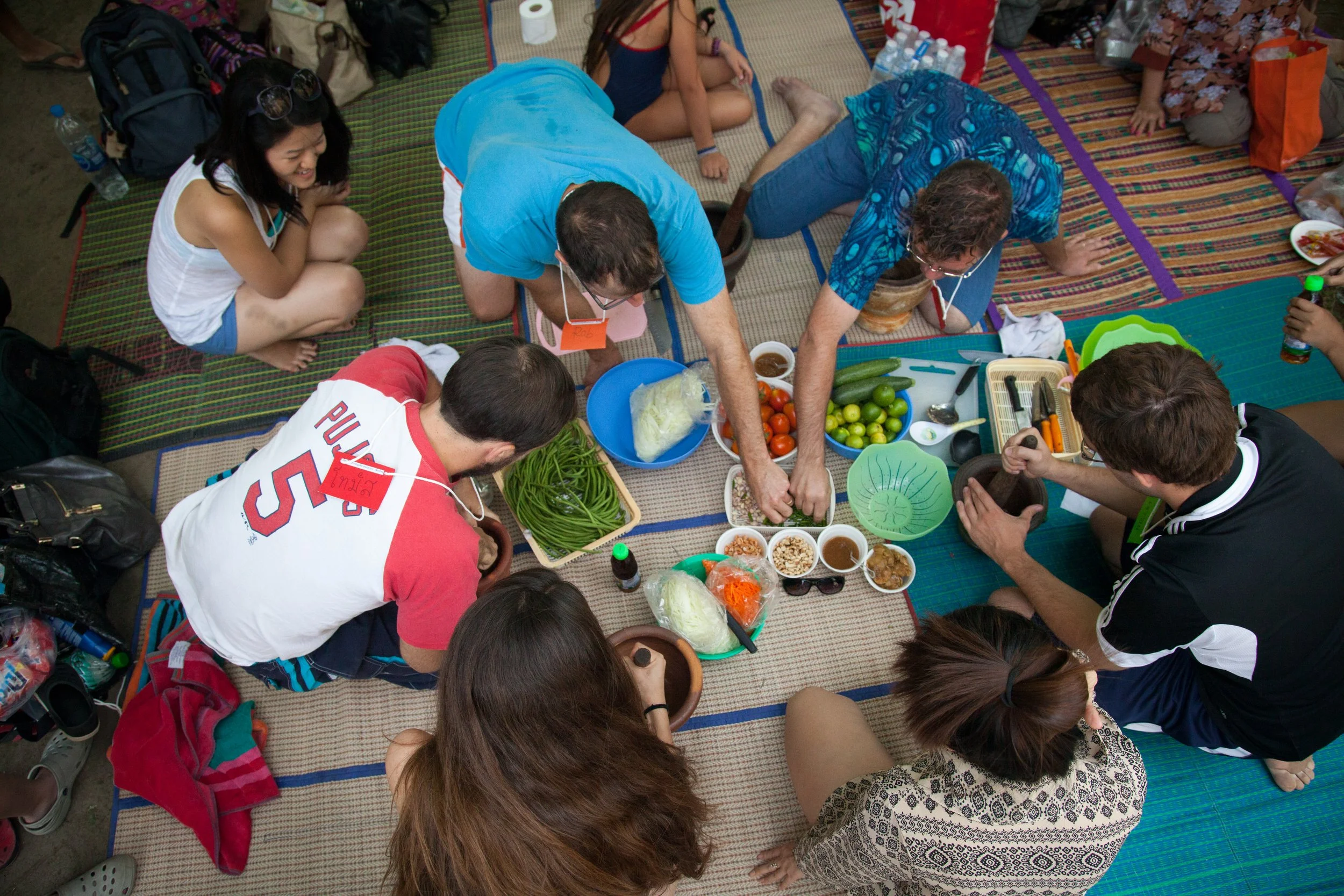 Group of people sitting on mats around a table, preparing food with vegetables and utensils
