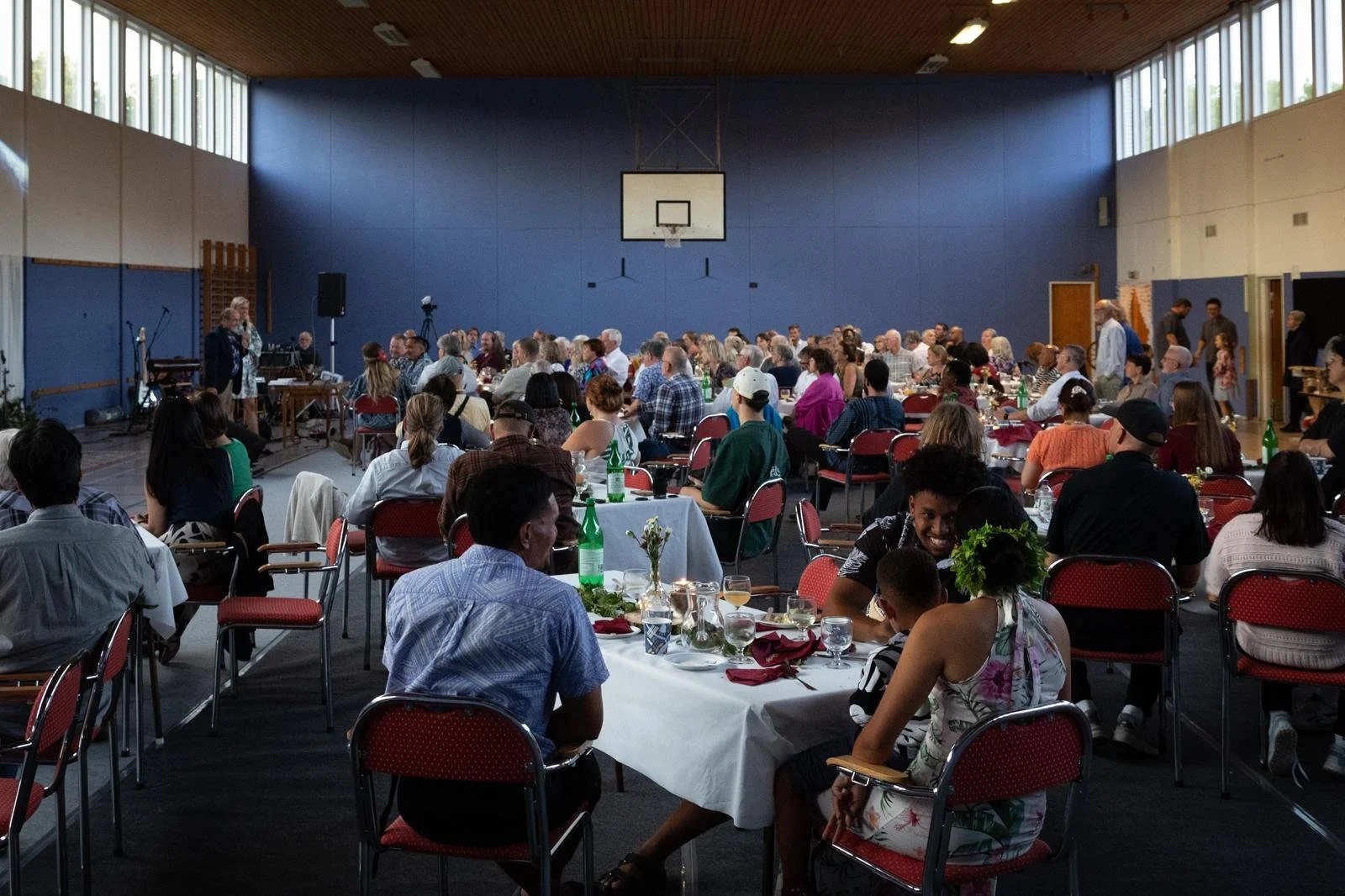 A large group of people sitting at round tables inside a gymnasium or hall, listening to a woman speaking at a microphone on a small stage, with musical instruments nearby, and a basketball hoop on the back wall.