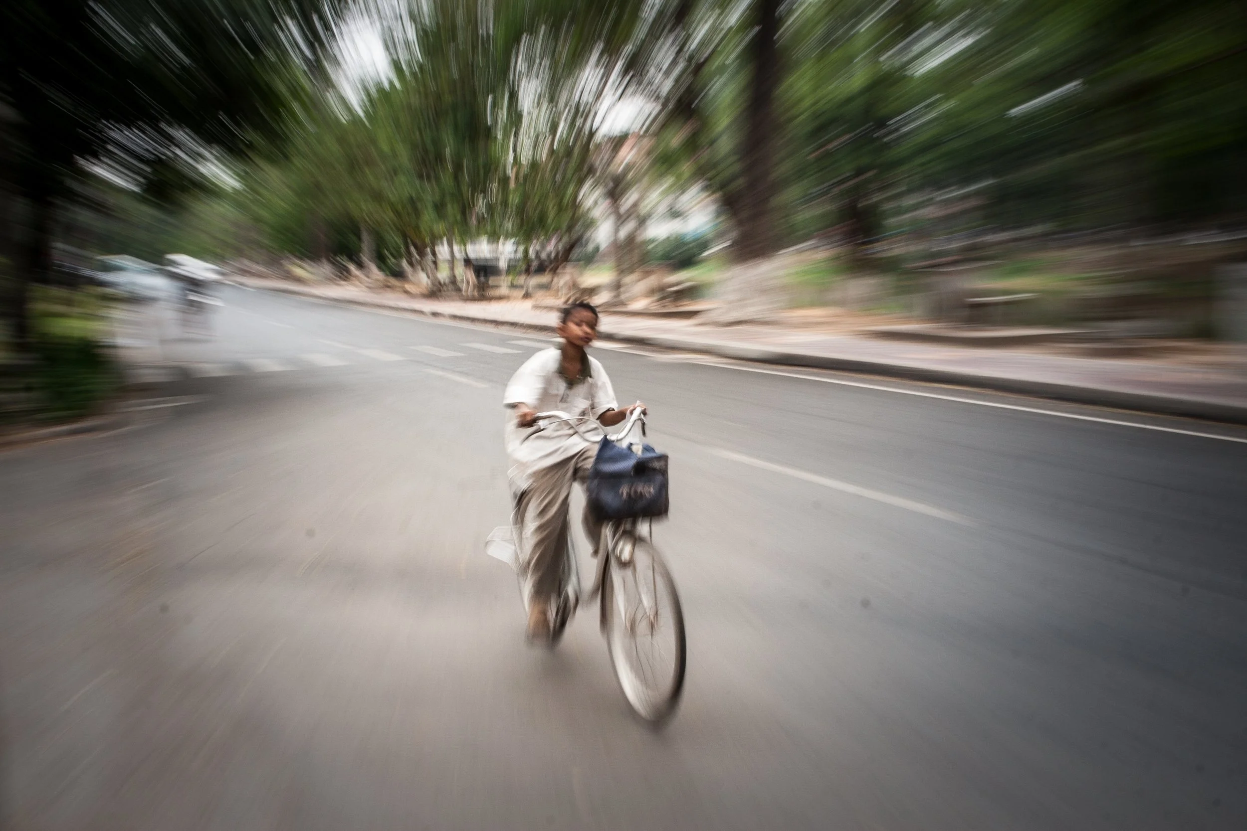 A woman riding a bicycle on a city street with trees in the background, motion blur indicating speed.