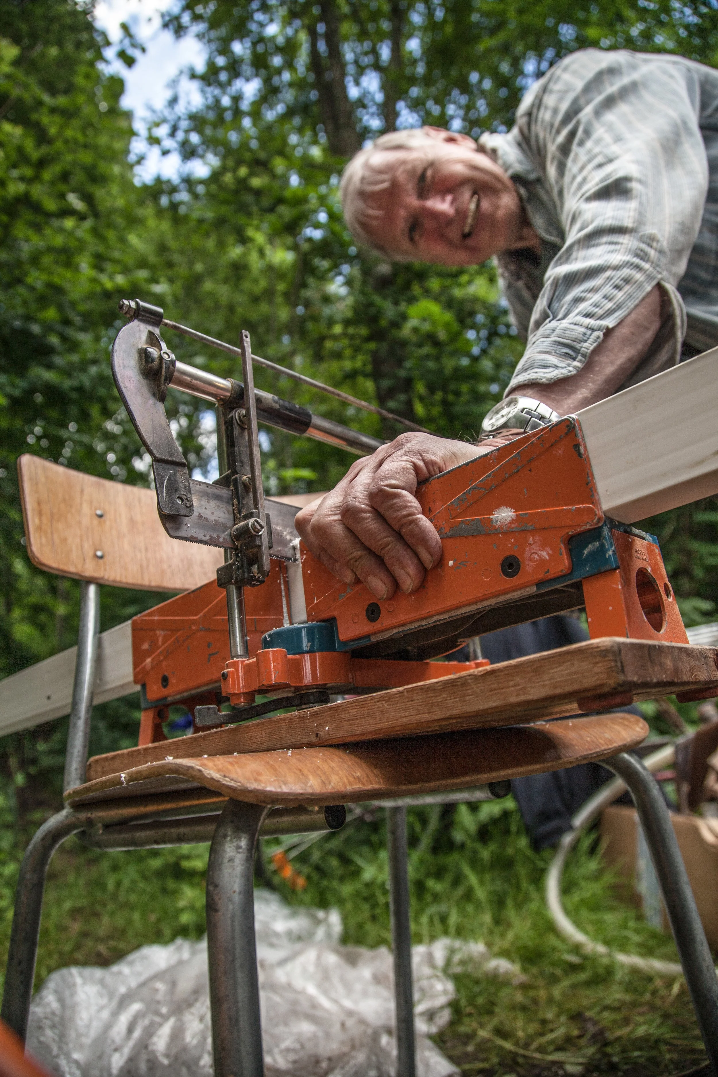 An elderly man with gray hair smiling while working on a woodworking project outdoors, using a hand saw on a piece of wood placed on a rustic wooden chair, with green trees in the background.