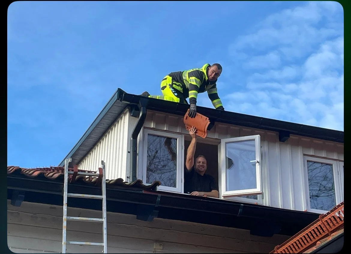 A firefighter in safety gear on the roof of a house handing a plastic container to a man at an open window below.