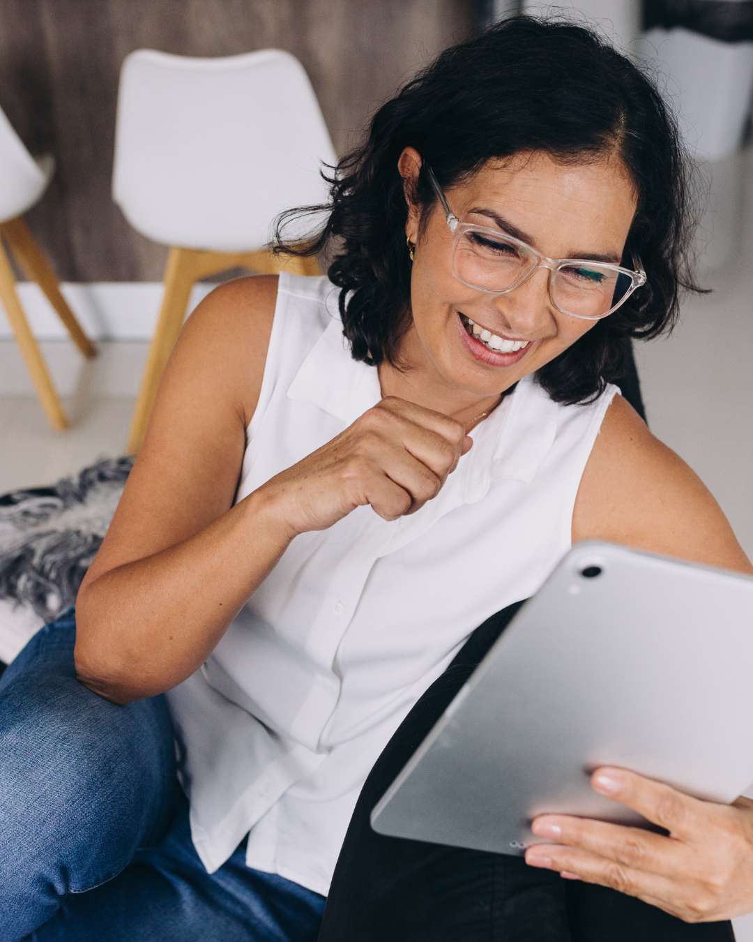 A woman with glasses, dark hair, and a white sleeveless blouse smiling and looking at a tablet.