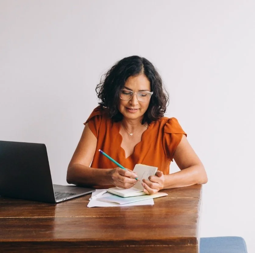 A woman with dark hair and glasses sitting at a wooden desk, writing in a notepad with a pen, with a laptop and some papers on the desk, against a plain white wall background.