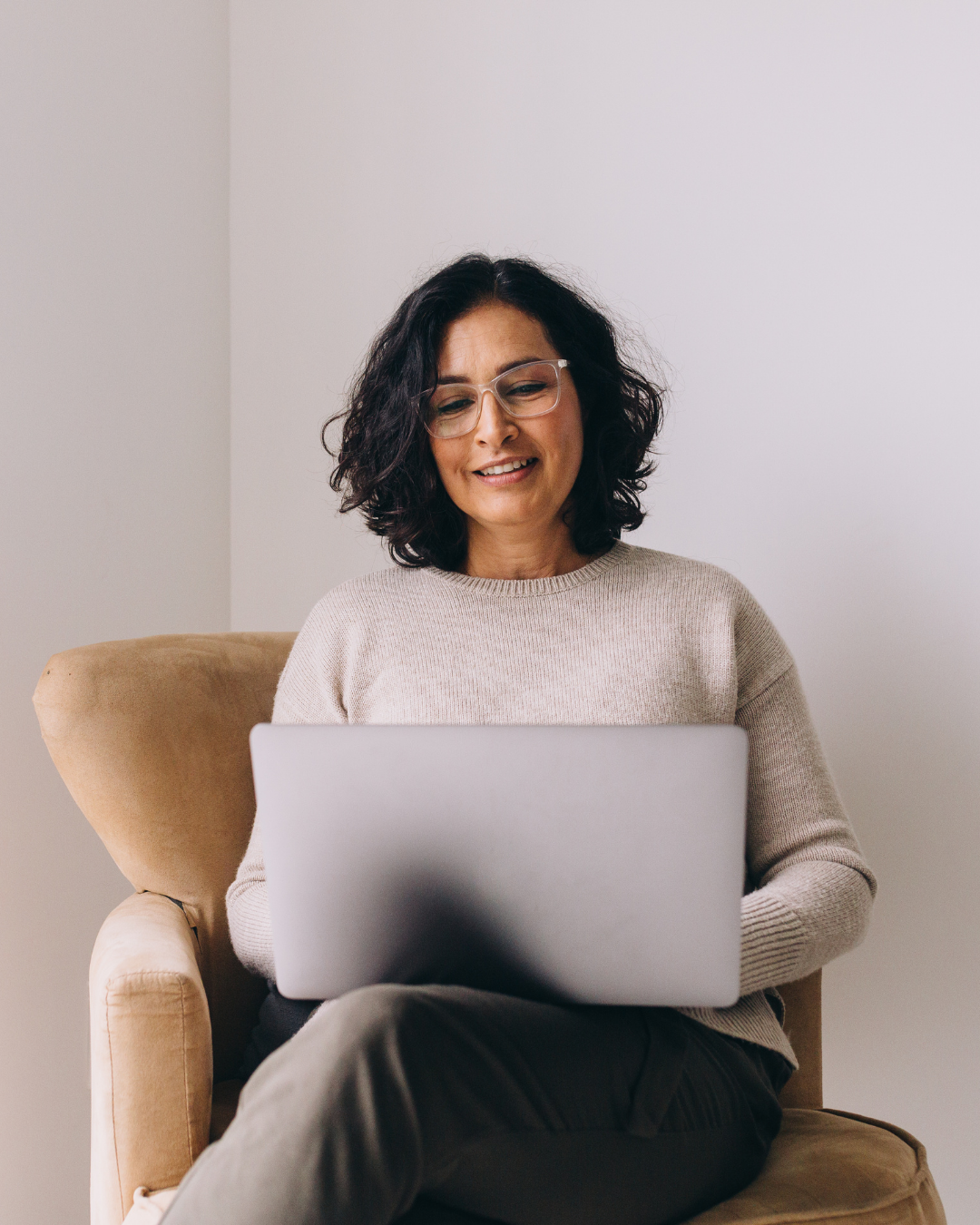 A woman with curly dark hair, wearing glasses and a beige sweater, sitting in a tan armchair, looking at her laptop with a slight smile.