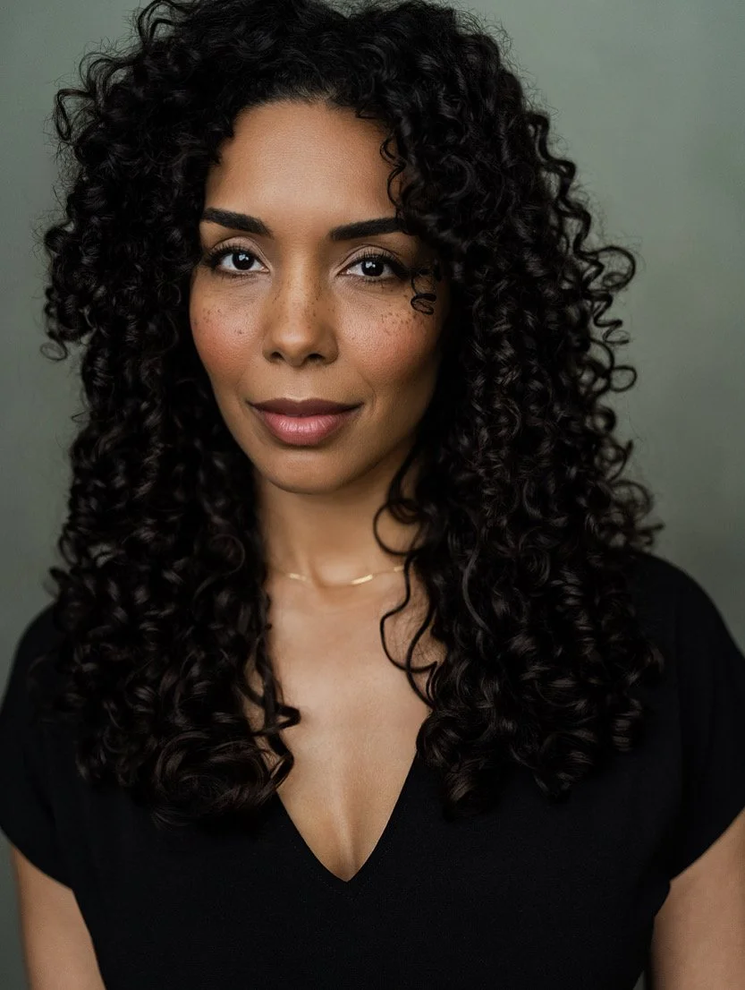 Close-up portrait of a woman with dark, curly hair, wearing a black top, smiling slightly, against a neutral background.