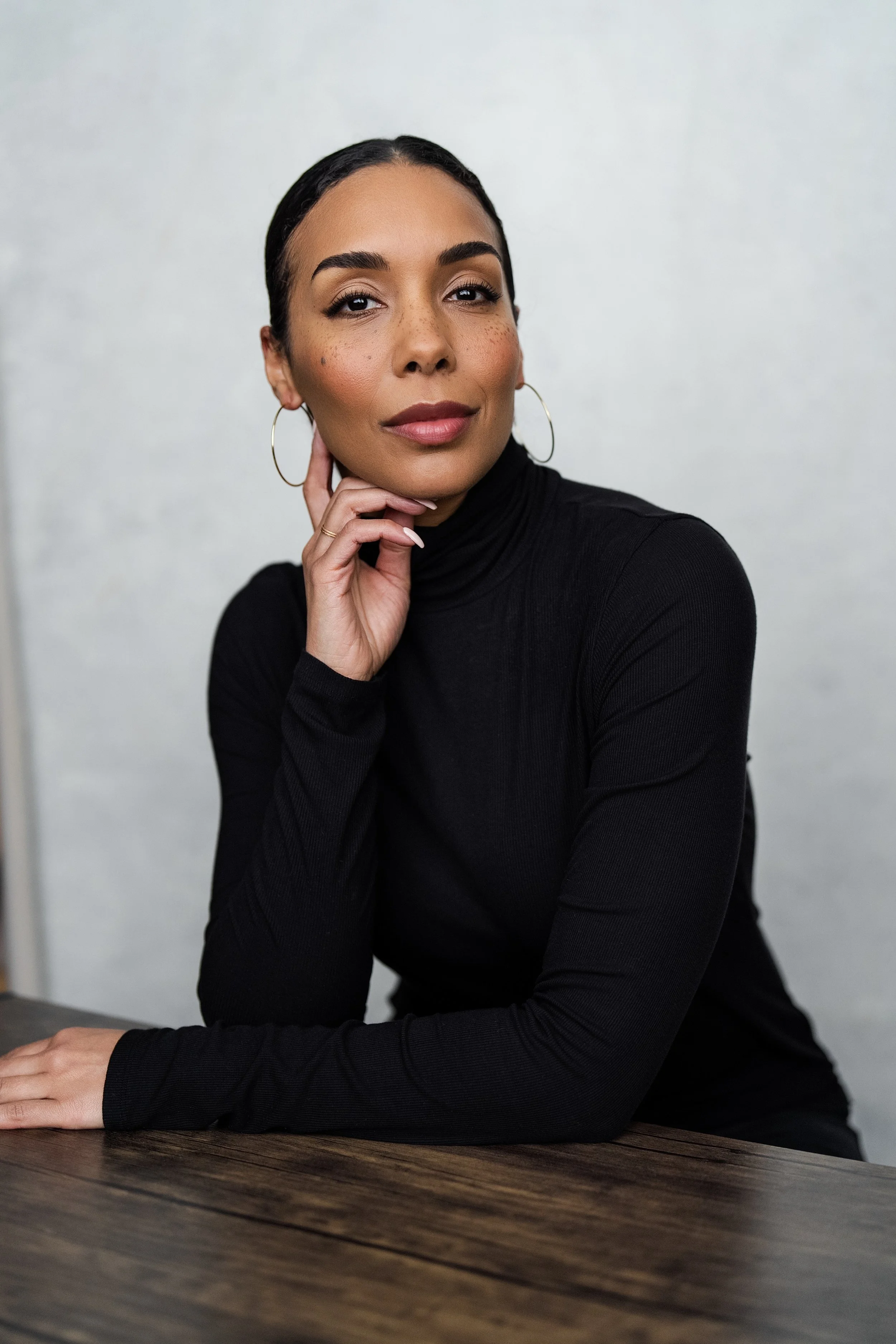 A woman with dark hair in a bun, wearing hoop earrings and a black turtleneck, sitting at a wooden table with her head resting on her hand, looking at the camera.