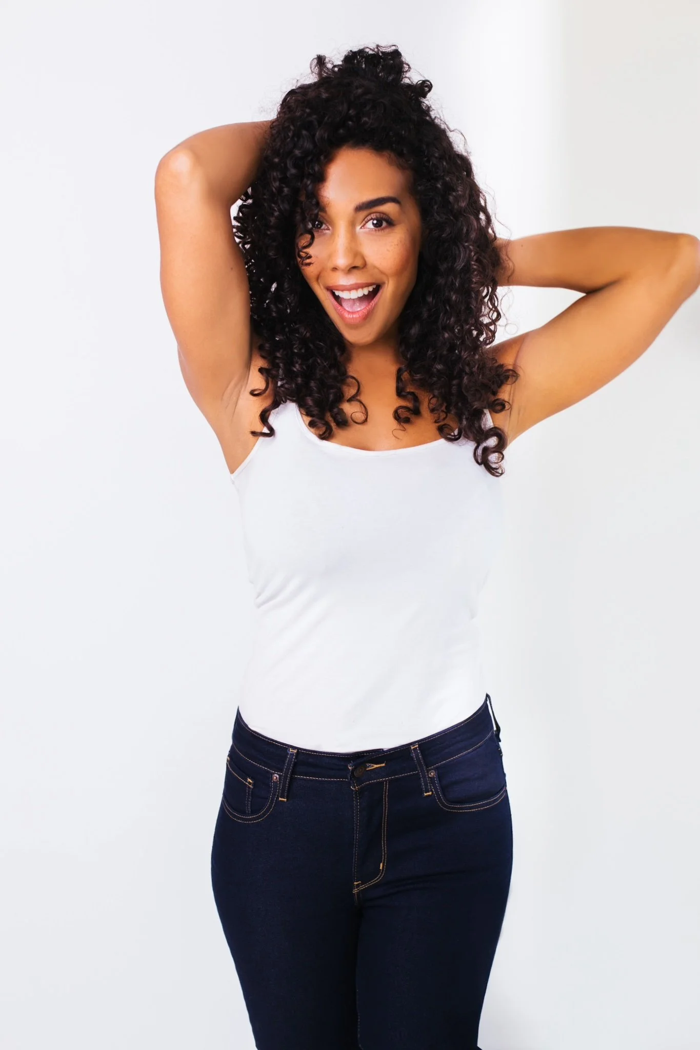 A woman with curly hair smiling and posing with her hands behind her head, wearing a white tank top and dark jeans.