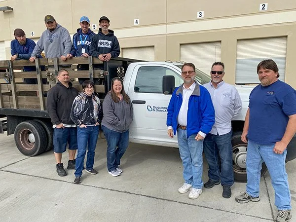 Group of nine people standing in front of a white truck with a flatbed loaded with wooden pallets. The truck has a logo that says 'Dugmore & Duncan'. The group is outside a beige warehouse building with numbered garage doors.