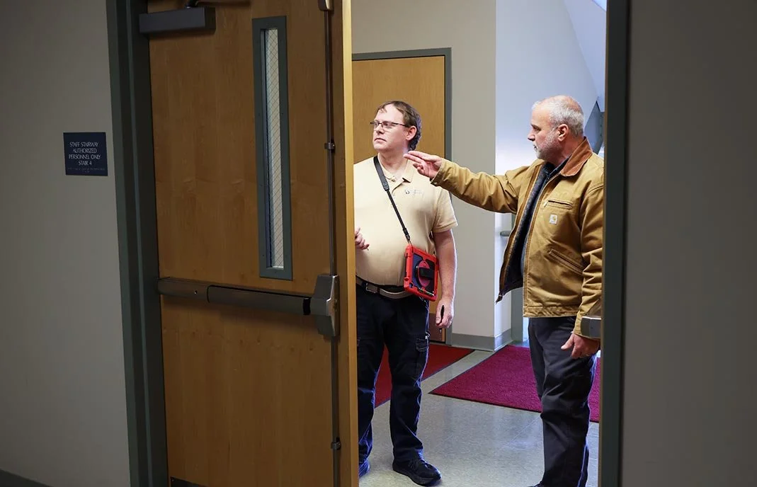 Two men standing near an open door in a hallway, engaged in conversation. One man is gesturing with his hand, and both are watching something outside the door.