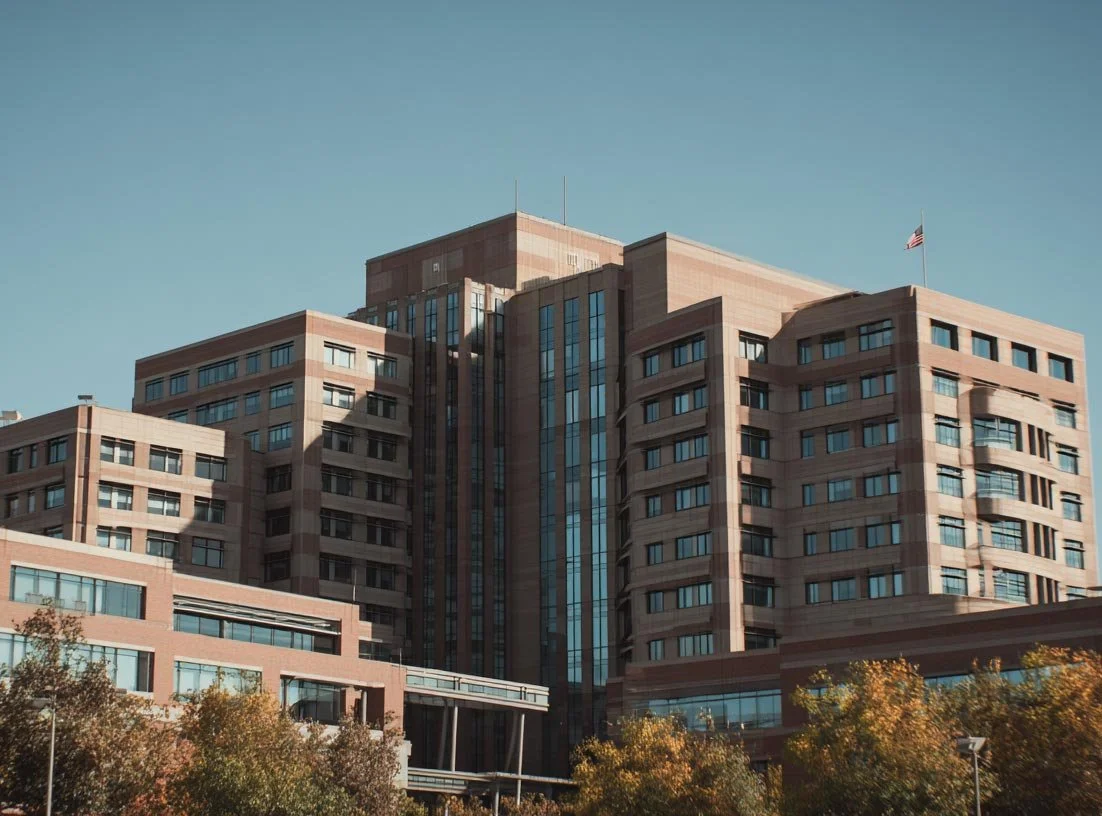 A large multi-story office building with a central glass tower, brick facade, and an American flag on the top, set against a clear blue sky.