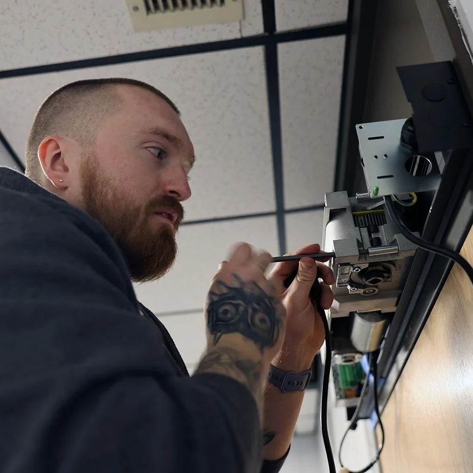 A man with a beard and tattoos working on electronic equipment mounted on a wall ceiling, using a screwdriver.
