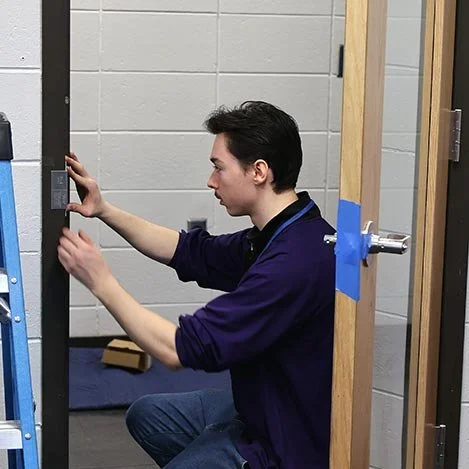 A young man is kneeling and working with a keypad on a metal structure, possibly assembling or repairing it in a workshop or gym setting.