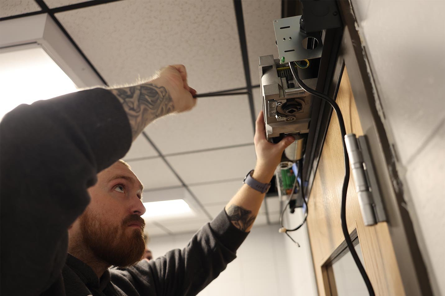 A man with tattoos on his arms is repairing or installing a device on a ceiling, using a screwdriver. The ceiling has a grid pattern with ceiling tiles, and the room features fluorescent lighting.