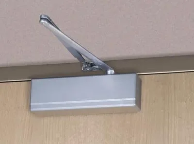 A door closer mounted at the top corner of a wooden door, with a metal arm extending from the door frame to the door.