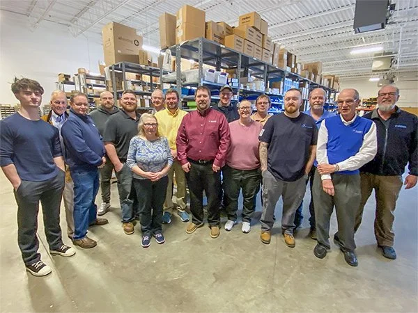 Group of 15 people standing in a warehouse aisle with shelves and boxes behind them.