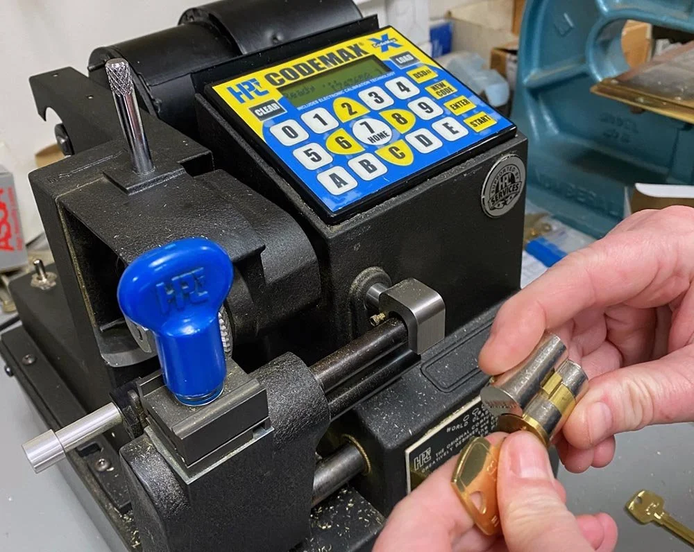 A hand holding a brass key and a brass lock cylinder on a key cutting machine in a workshop.