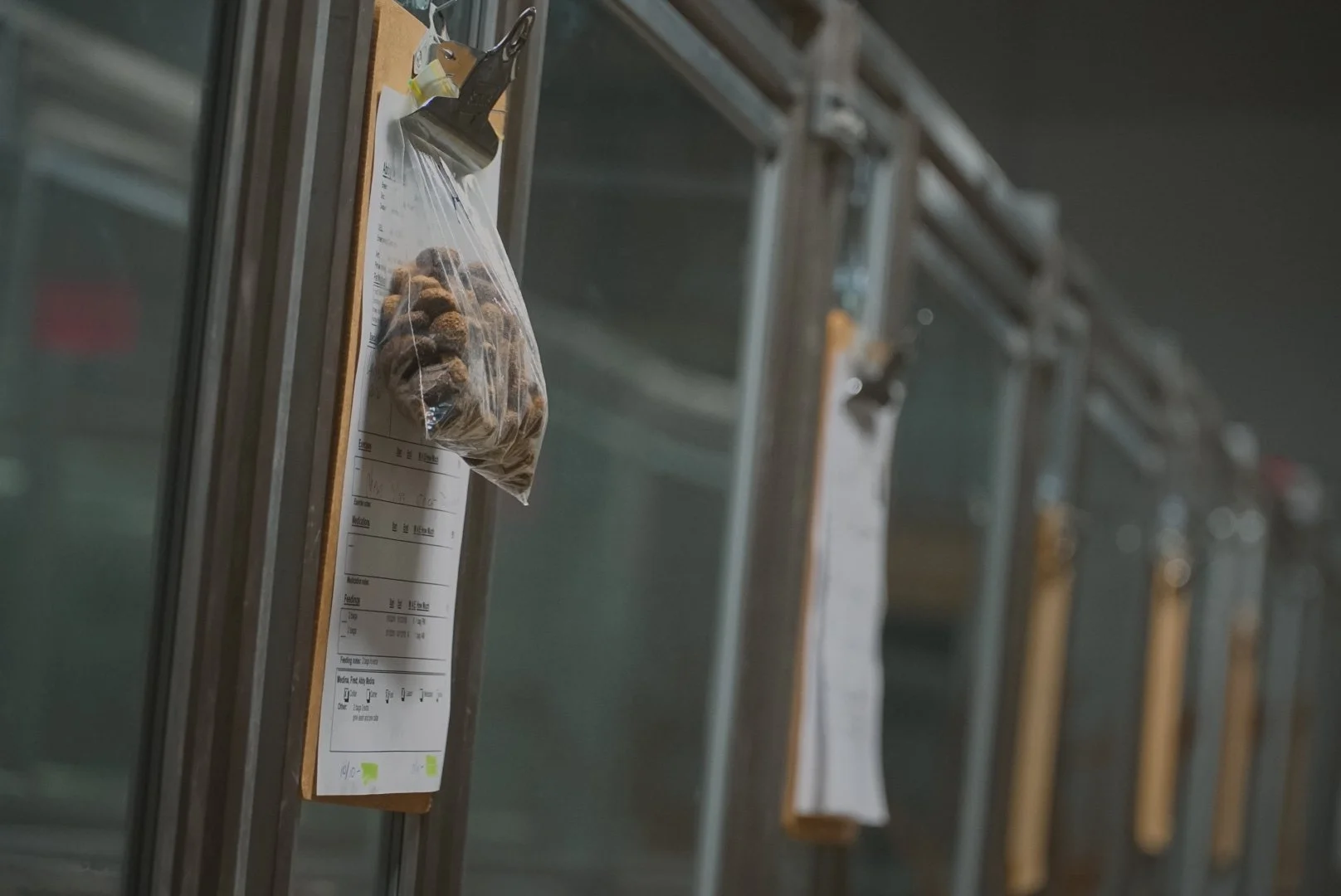 A clipboard with a plastic bag of what appears to be cinnamon rolls or baked goods hanging from it. Multiple similar clipboards are attached to rails or dividers in the background.
