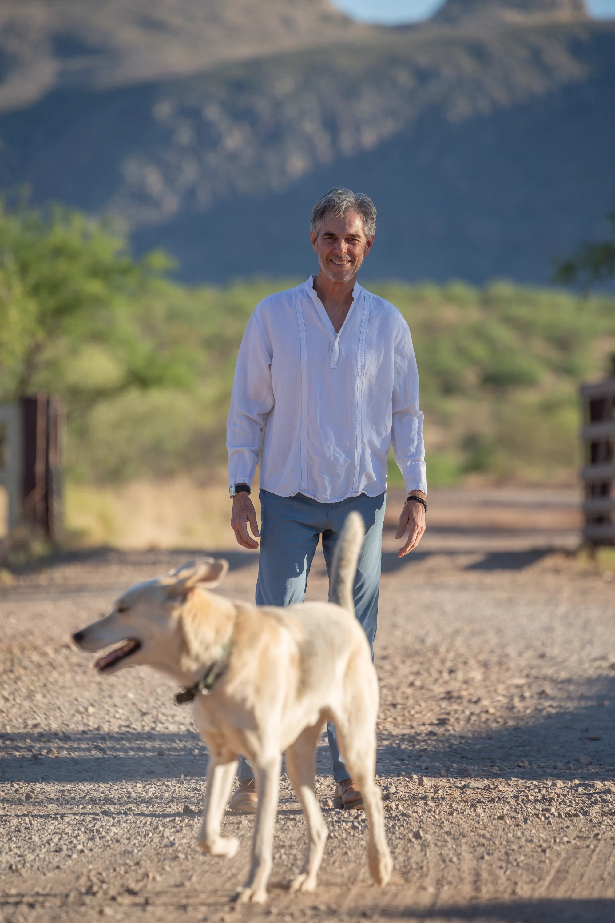 A man with gray hair smiling, walking outdoors with a light-colored dog on a dirt path, greenery and mountain in the background.