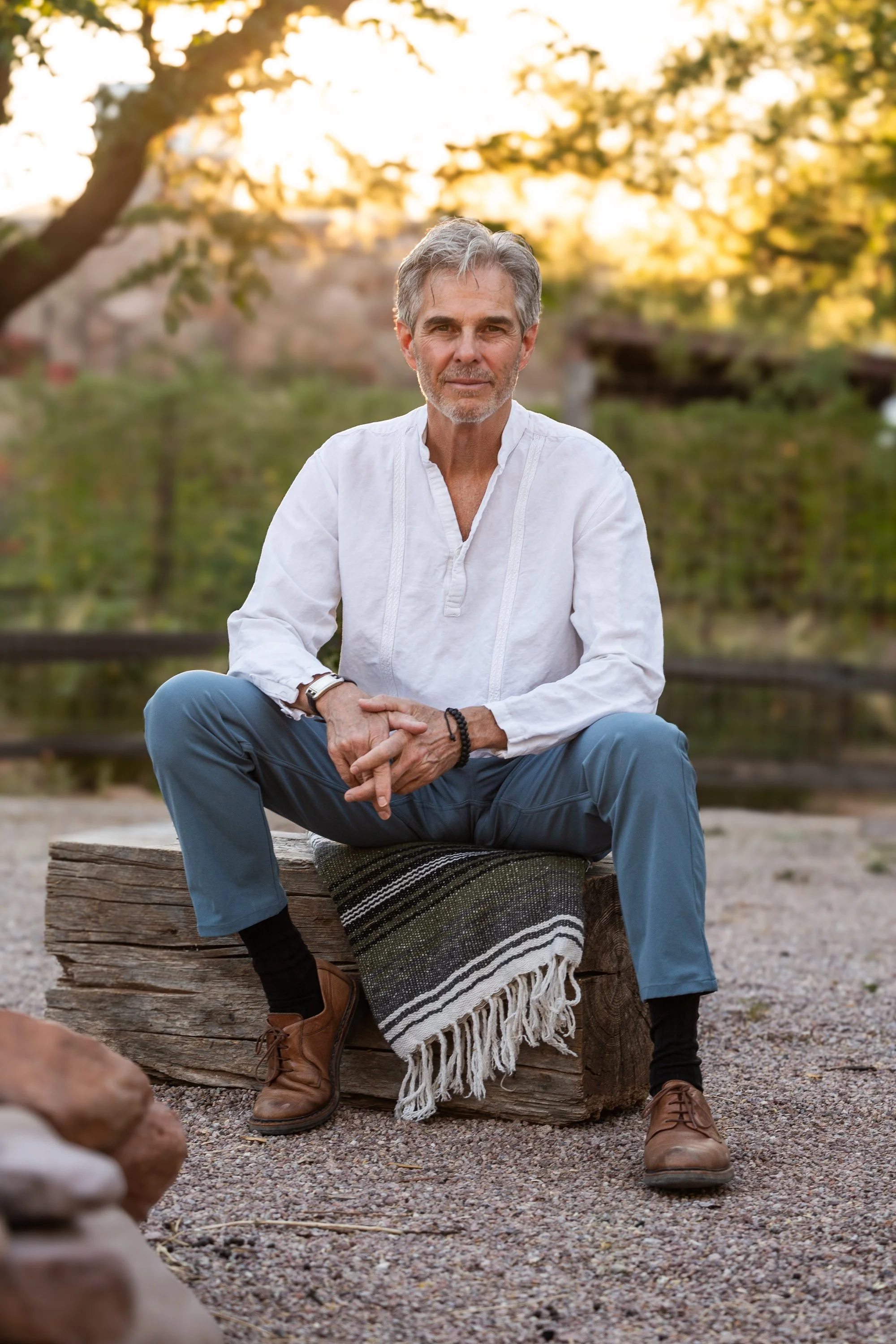 A middle-aged man with gray hair and a beard is sitting outdoors on a rustic wooden bench with a woven blanket draped over it. He's wearing a white linen shirt and blue trousers, with brown shoes and black socks. The background reveals a sunset with trees and greenery, creating a warm, serene atmosphere.