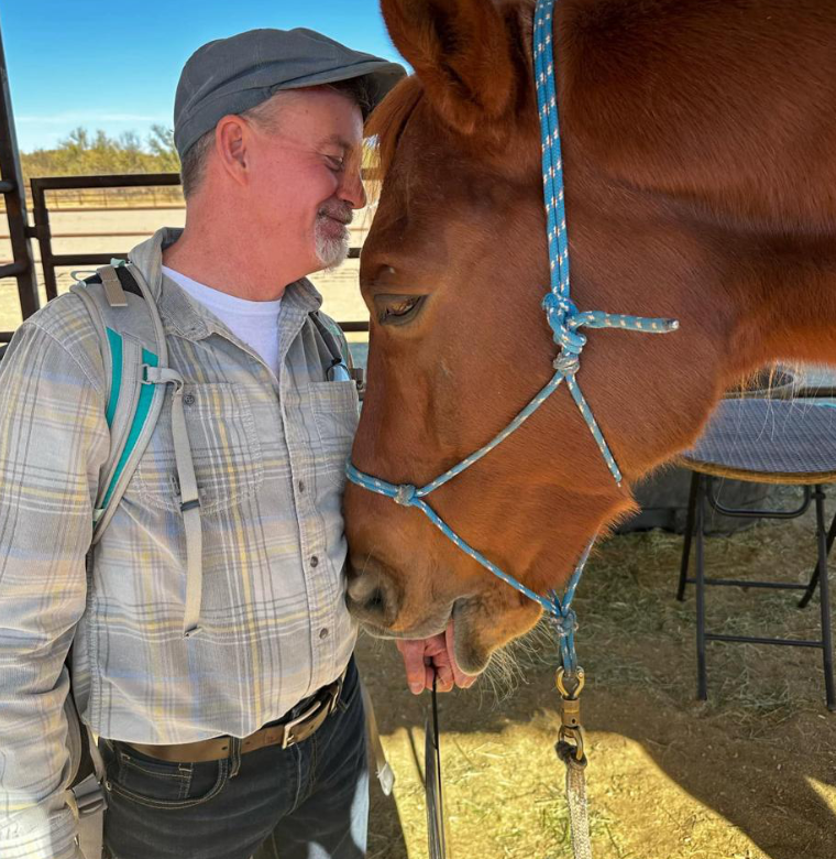 A man wearing a gray hat and plaid shirt touching foreheads with a brown horse, both with closed eyes, in an outdoor setting with a fence and blue sky in the background.