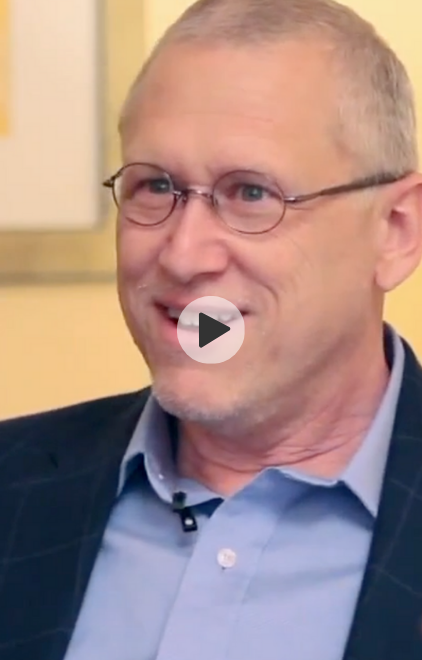 A middle-aged man with short gray hair and glasses, smiling and speaking, wearing a suit jacket and a light blue shirt.