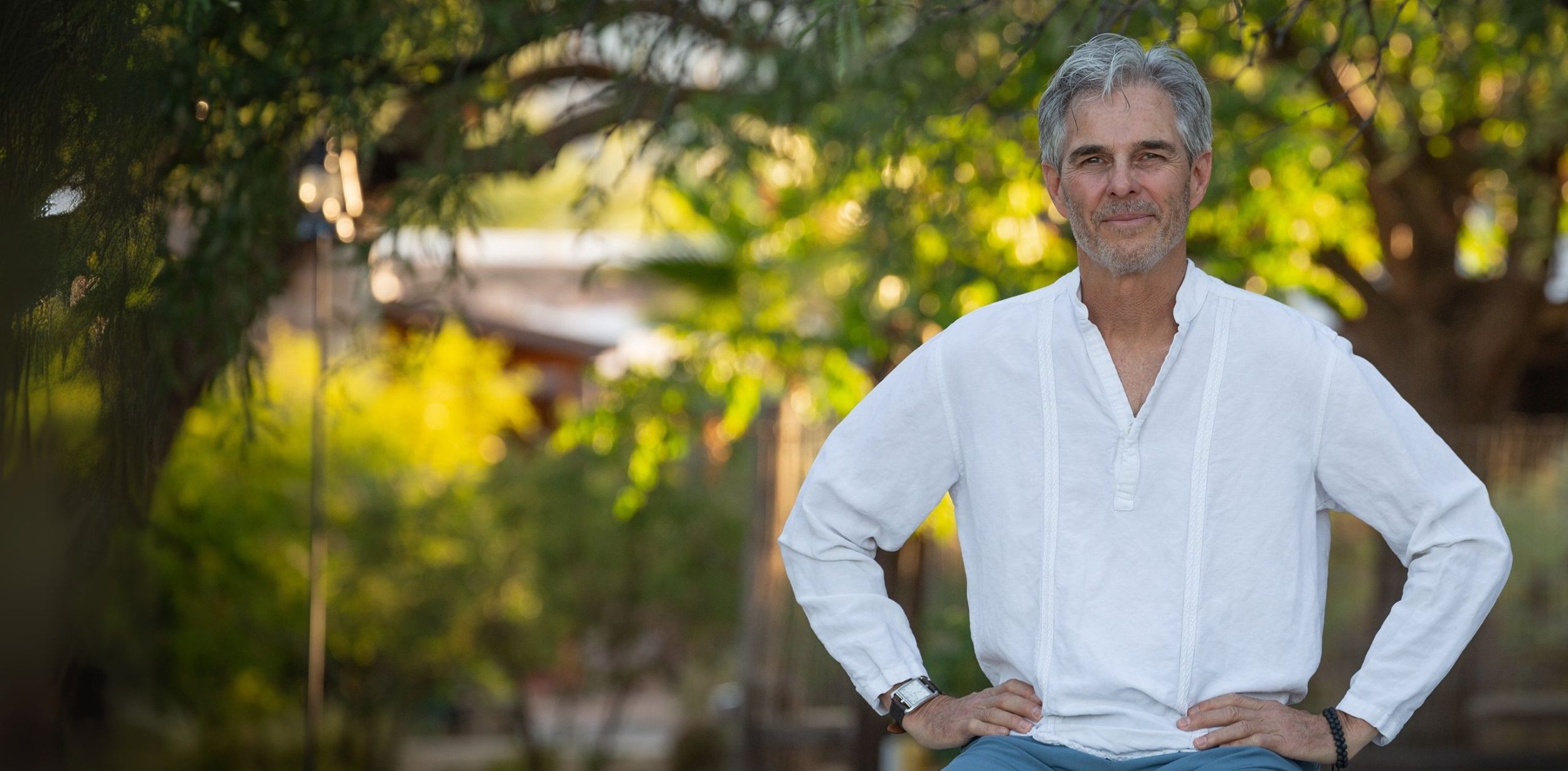 An older man with gray hair and a beard standing outdoors with his hands on his hips, wearing a white long-sleeve shirt, in a garden or park setting with green trees and sunlight.