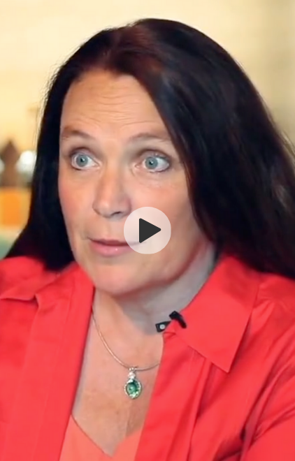 A woman with black hair and blue eyes wearing a red blouse and a green pendant necklace, speaking in an interview.