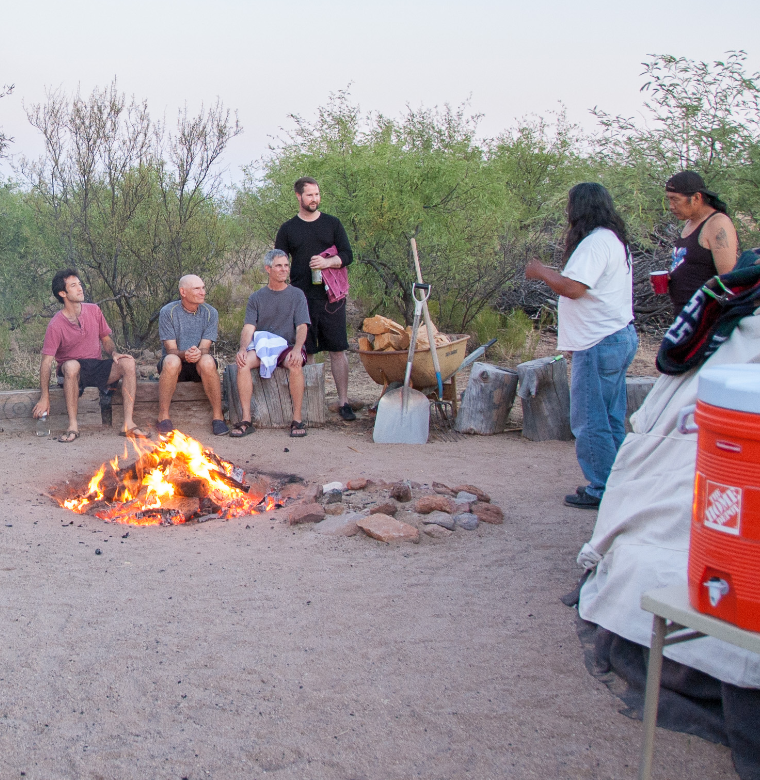 Group of people gathered around a campfire outdoors during dusk, with some sitting on logs and others standing, surrounded by trees and camping gear.