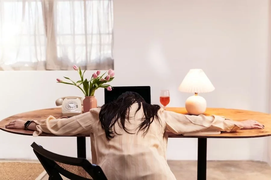 A woman with dark hair sitting at a wooden table with her arms outstretched, facing away from the camera, in a room with a window, a lamp, a computer, and a vase of pink tulips.