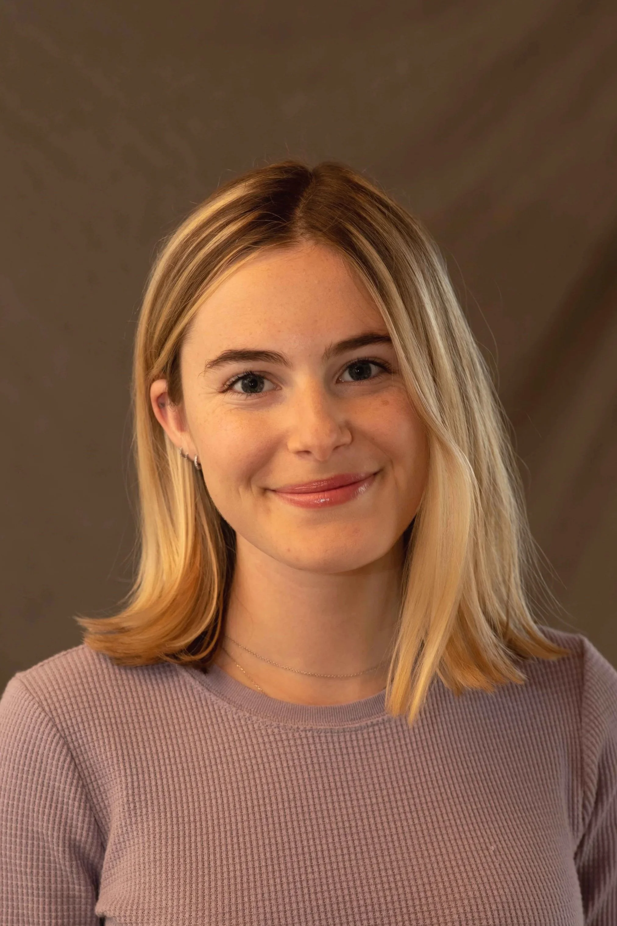 A young woman with blonde hair and a subtle smile, wearing a lavender top and small earrings, against a blurred brownish background.