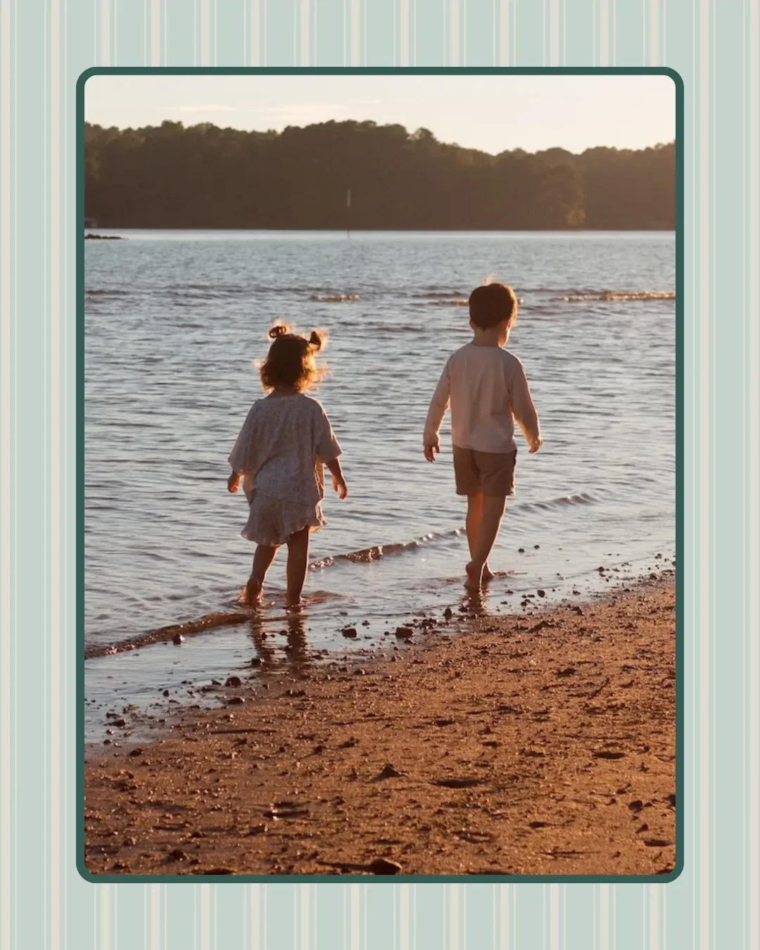 Two children, a girl and a boy, walk barefoot along a beach shoreline at sunset, with water and trees in the background.