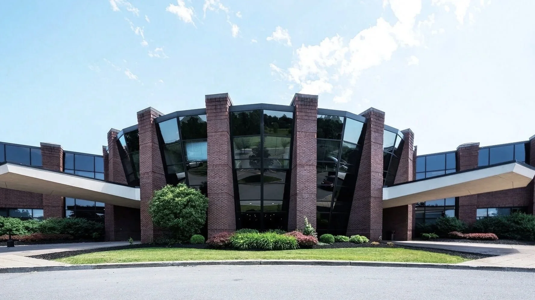Modern building with curved glass windows, brick columns, and a landscaped front yard with plants and bushes, under a partly cloudy sky.