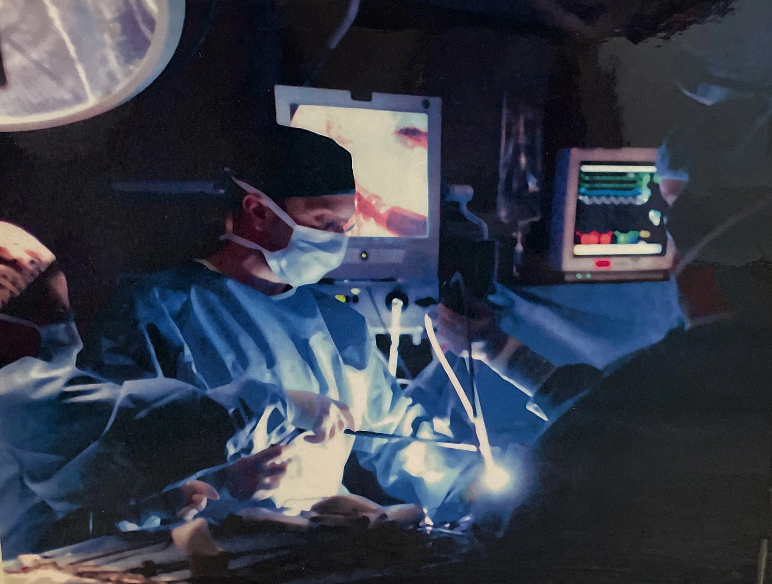 Dr. George Blessios and one other medical professional in surgical scrubs, wearing masks and gloves, performing a surgery in a darkened operating room with monitors displaying medical images.